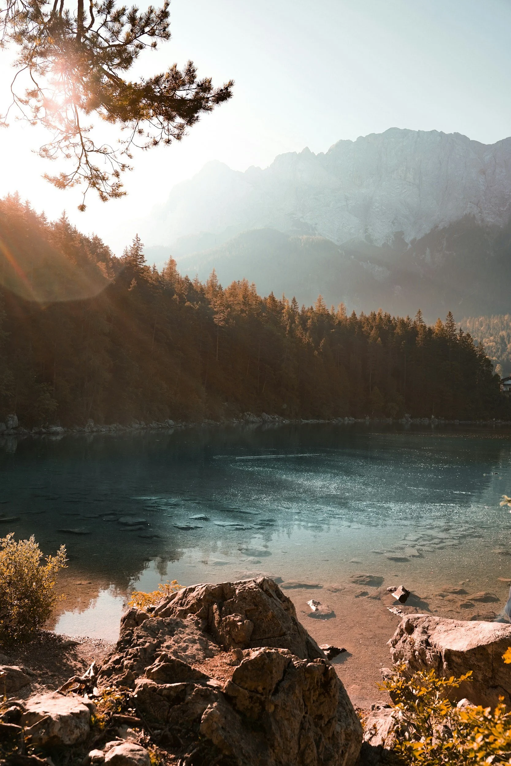 Scenic view of a lake with a forested shoreline, large rocks in the foreground, and mountains in the background, illuminated by sunlight.