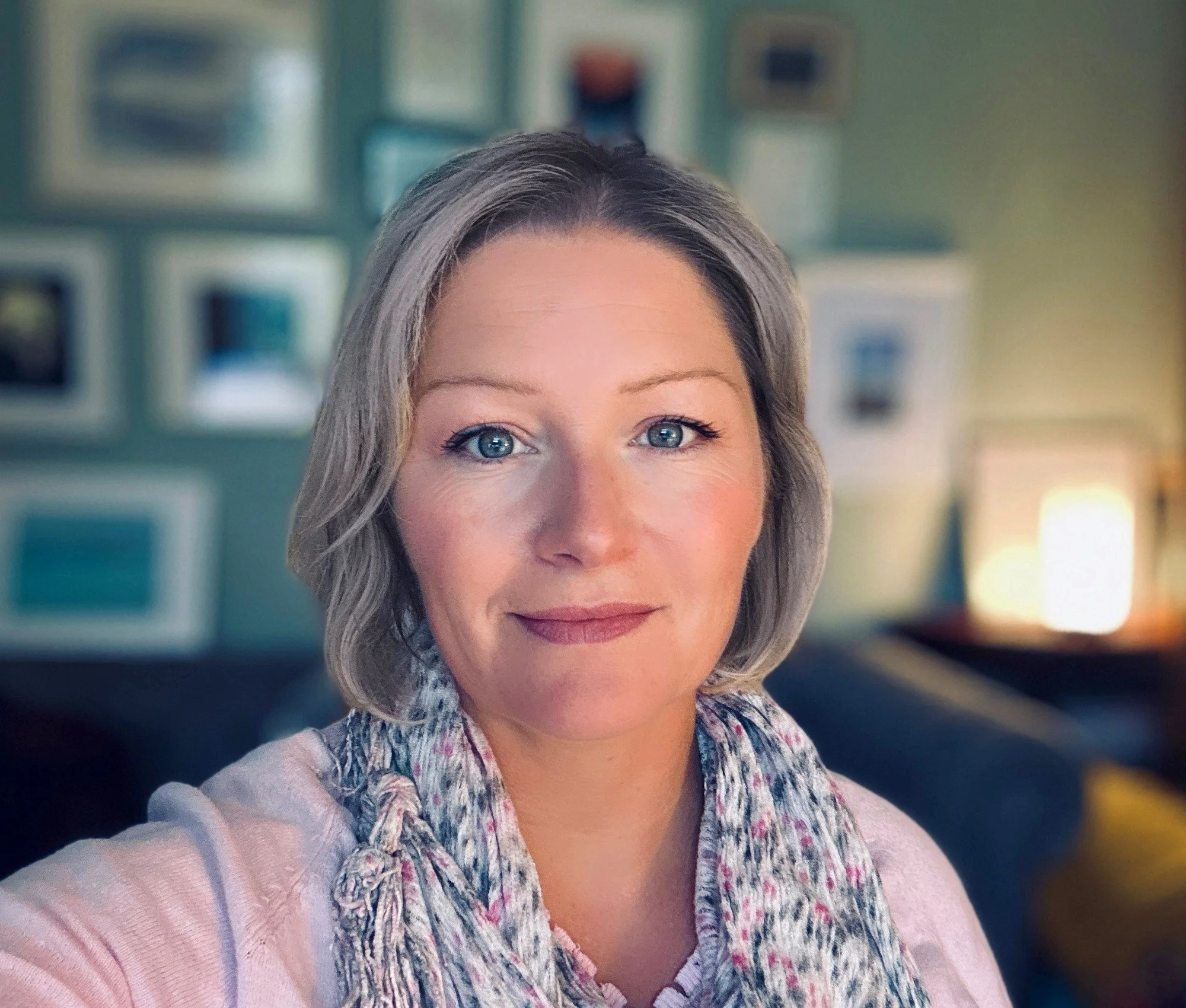 Close-up of a woman with light-colored hair and blue eyes, wearing a patterned scarf and pink sweater, taking a selfie in a room with shelves and decorations in the background.