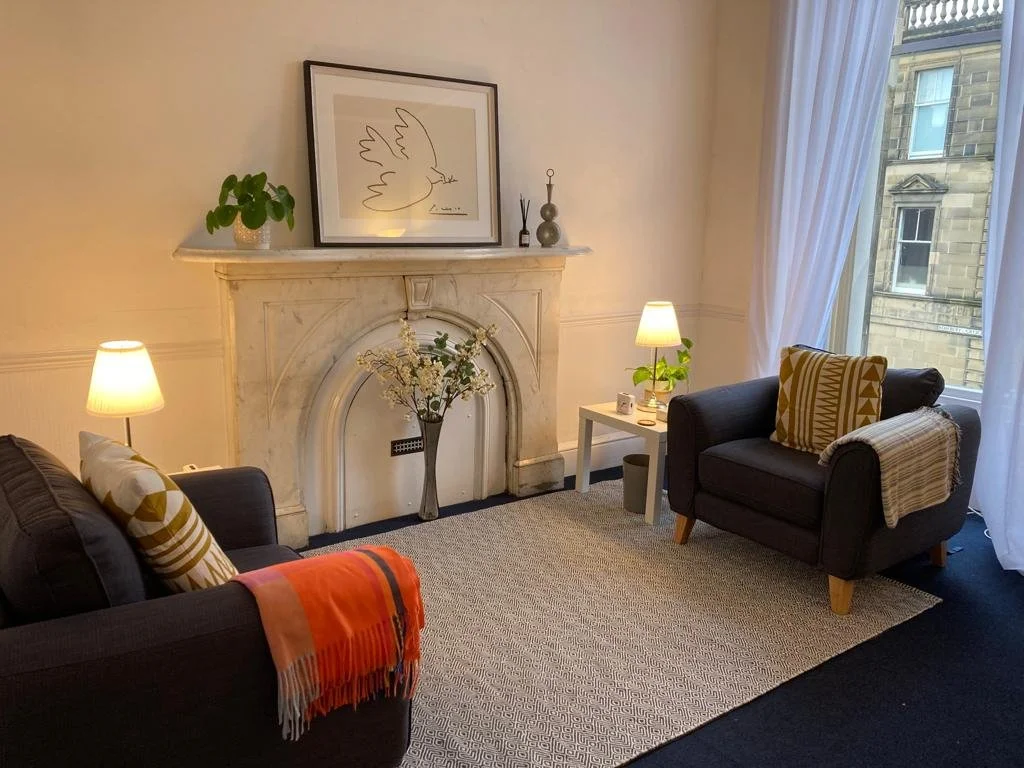 Living room with two black armchairs, each with a pattern pillow and throw blanket, a white side table with a small lamp and plant, a fireplace with a vase of flowers on top, and framed artwork of a dove above the fireplace. Large window with white curtains showing an exterior building.