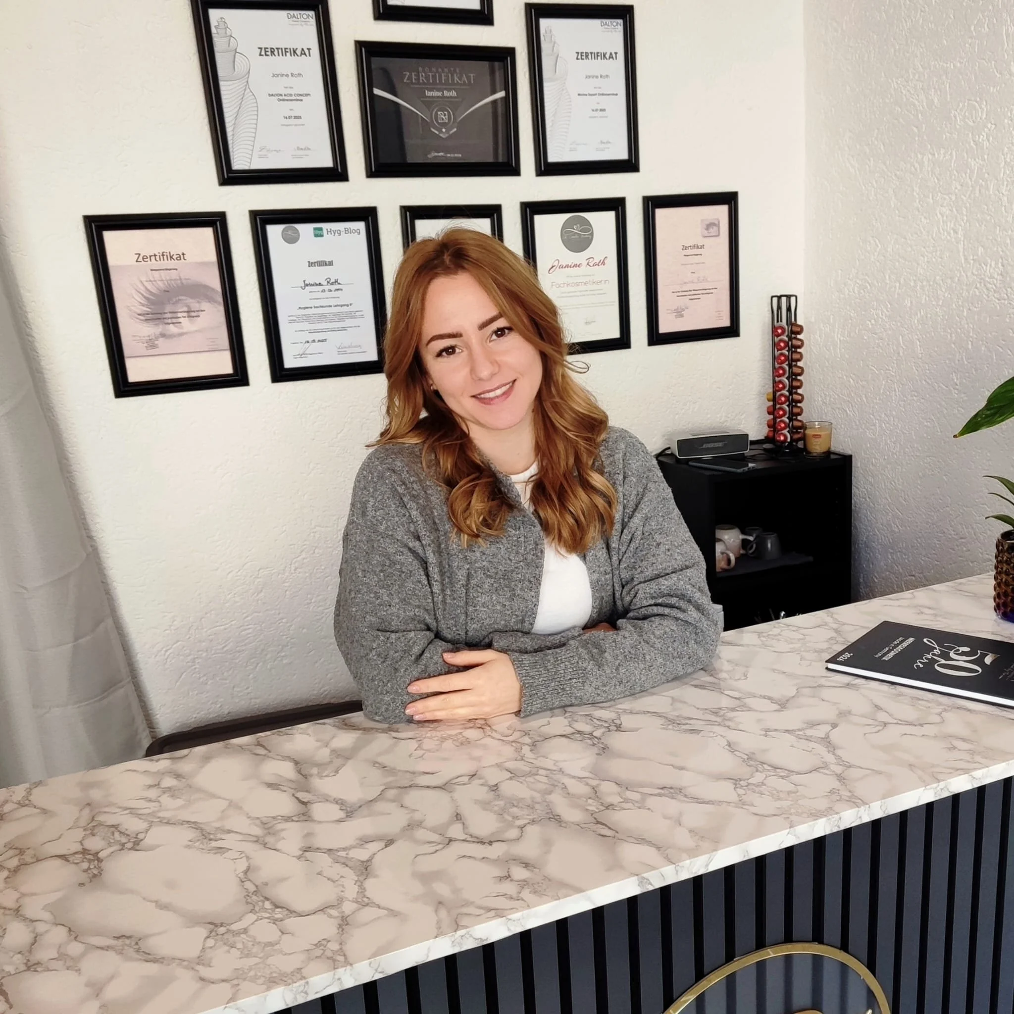 Eine Frau sitzt an einem Schreibtisch vor einer Wand mit mehreren eingerahmten Zertifikaten.