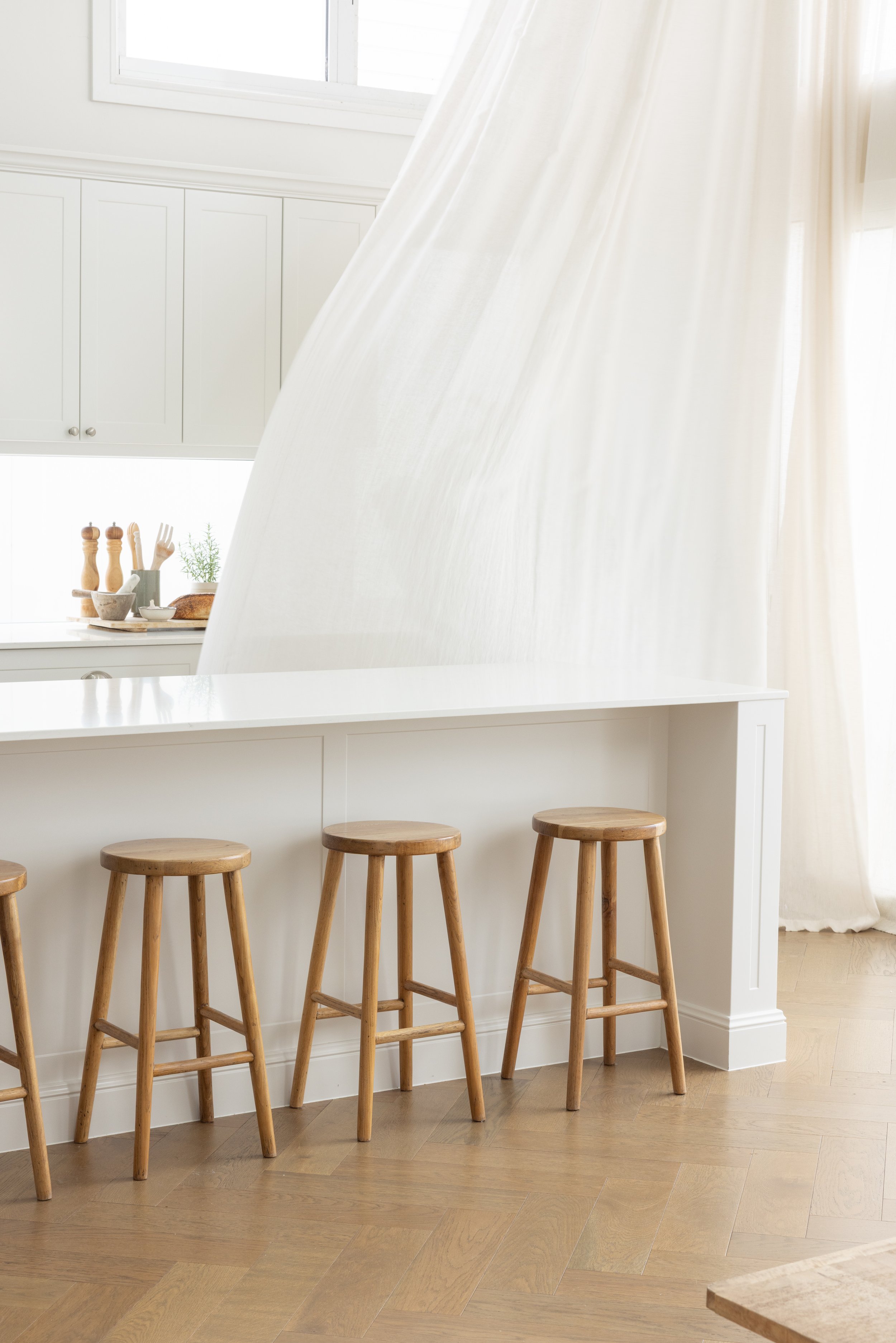 A white kitchen island with three wooden bar stools, a white countertop, and a light-colored wood floor. In the background, there are white kitchen cabinets, a window, and a small countertop with kitchen utensils, bread, and a small plant.