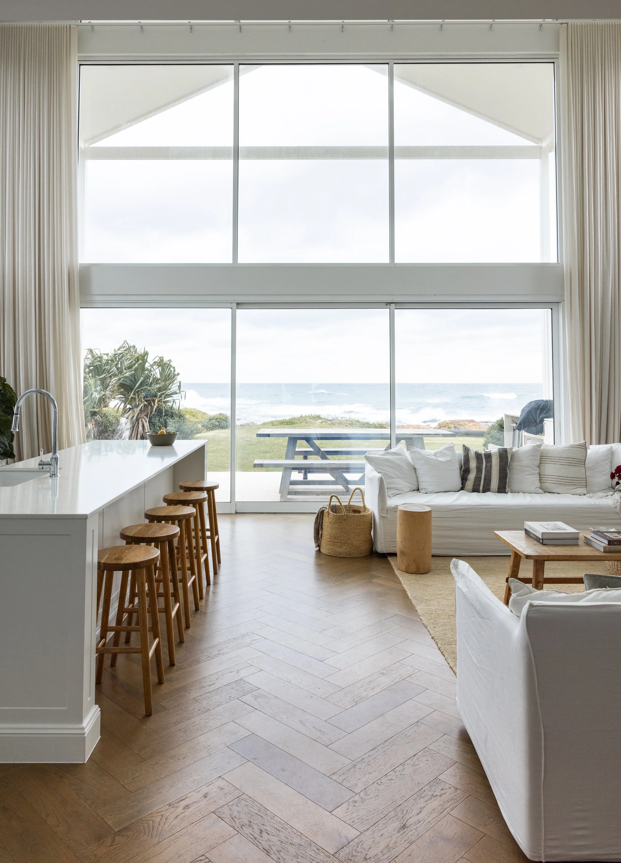 Living room with large windows overlooking a beach and ocean, wooden flooring, white furniture, and a kitchen area with bar stools.