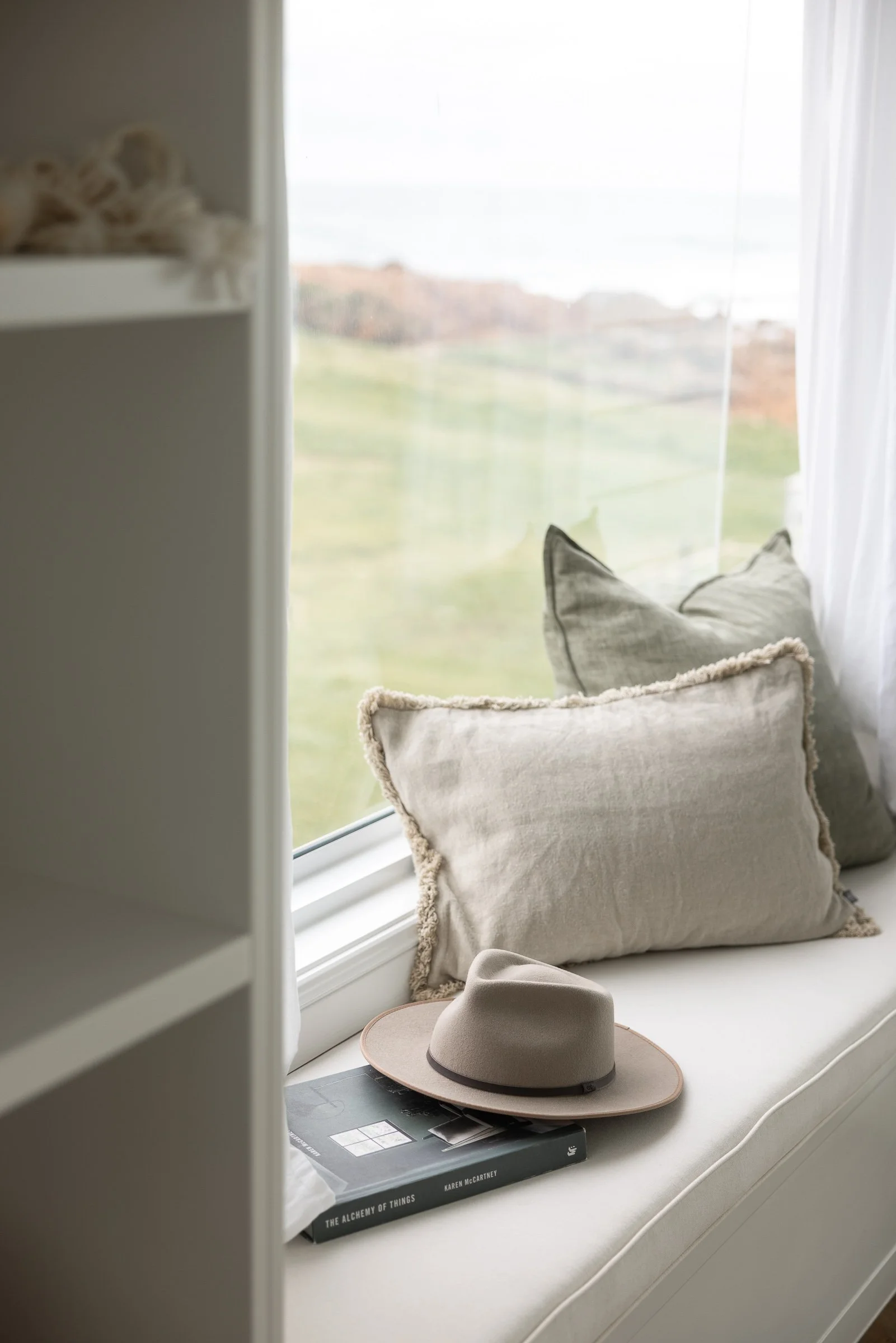 Window seat with beige pillows, a beige hat, and a book titled 'The Alchemy of Things' by Karen McCartney, overlooking a blurred green outdoor landscape.