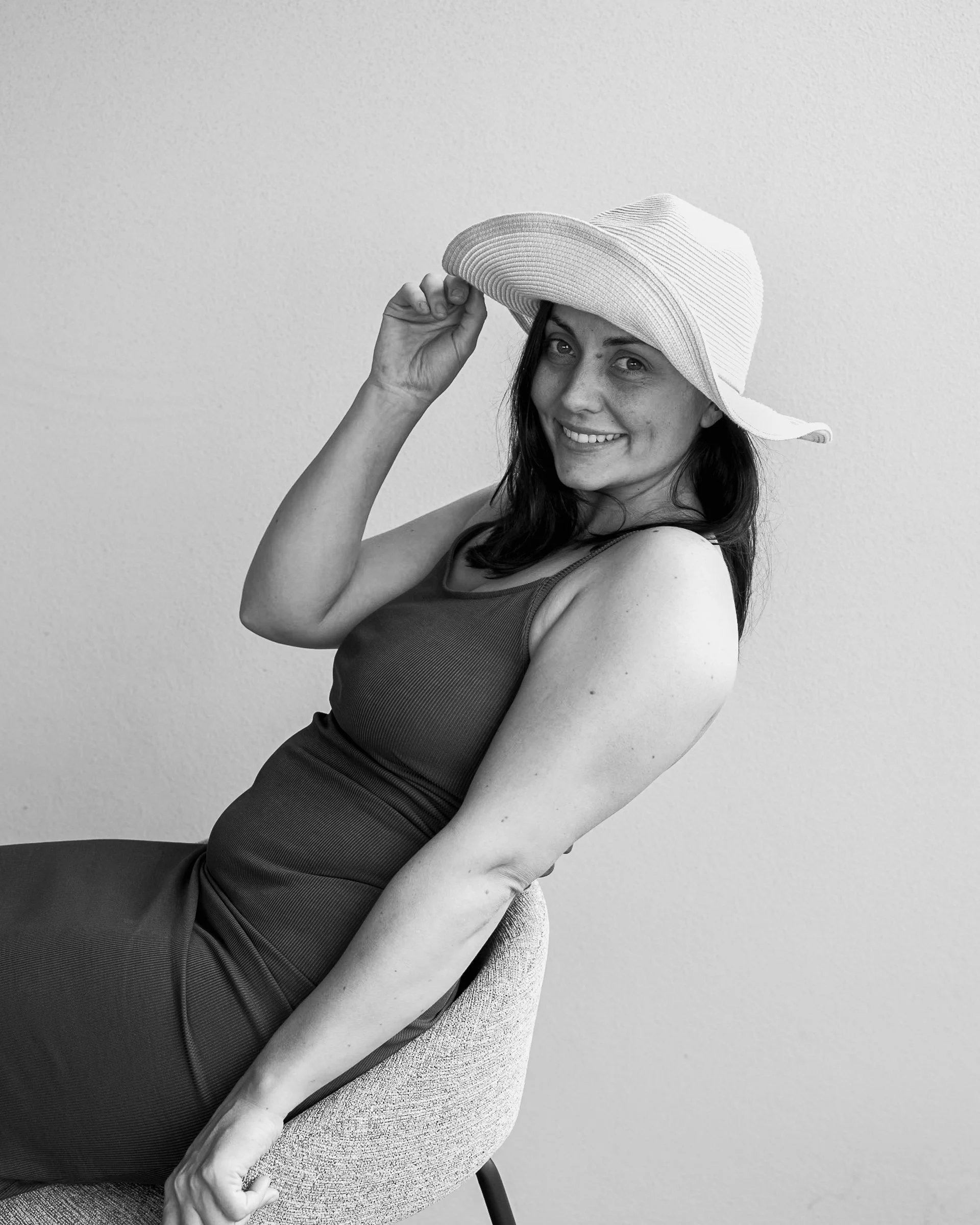 A woman sitting in a chair, smiling, wearing a sleeveless dress and a wide-brimmed hat, holding the brim of her hat with one hand, against a plain wall.