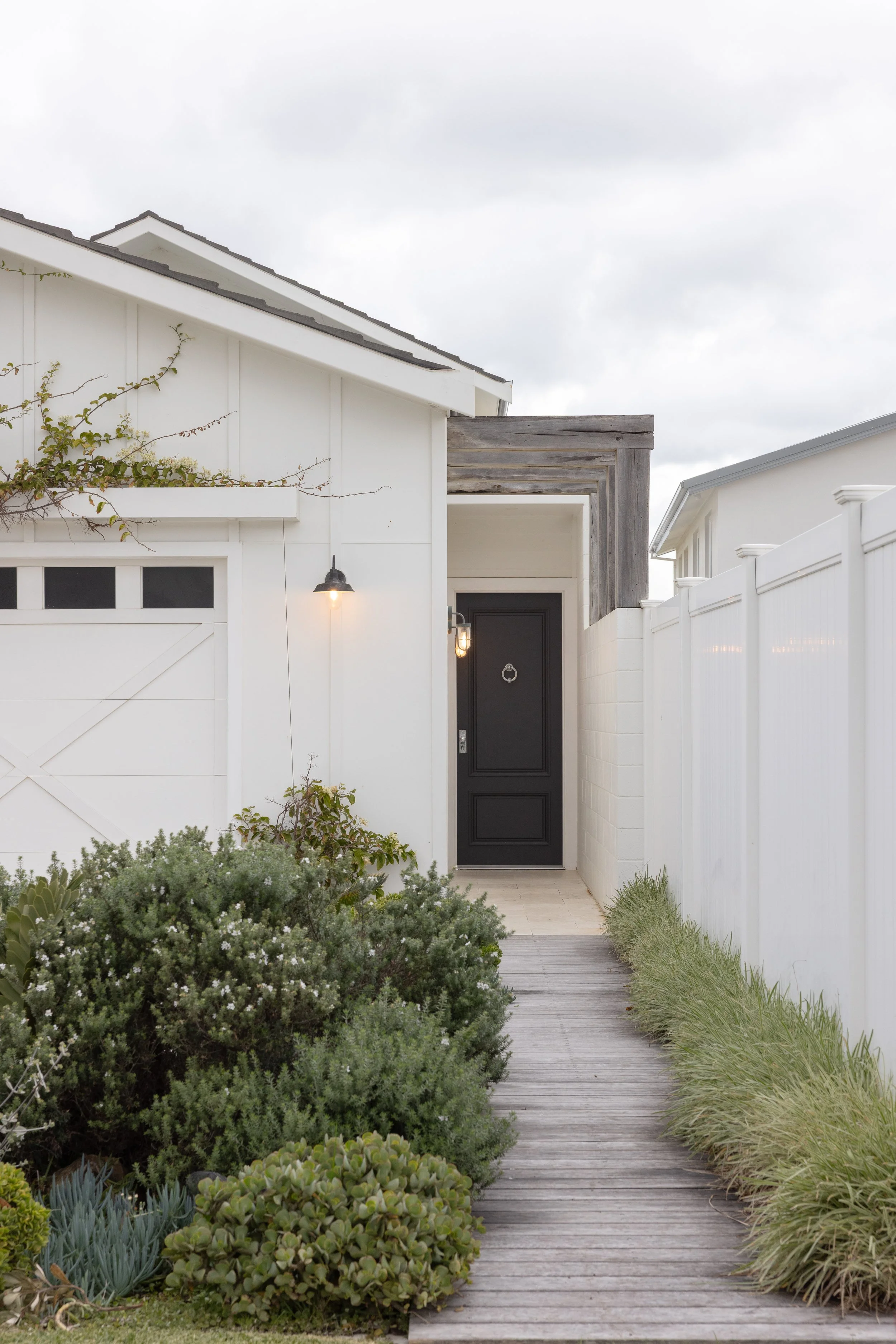 Front entrance of a house with a black door, white walls, wooden pathway, and garden bushes.