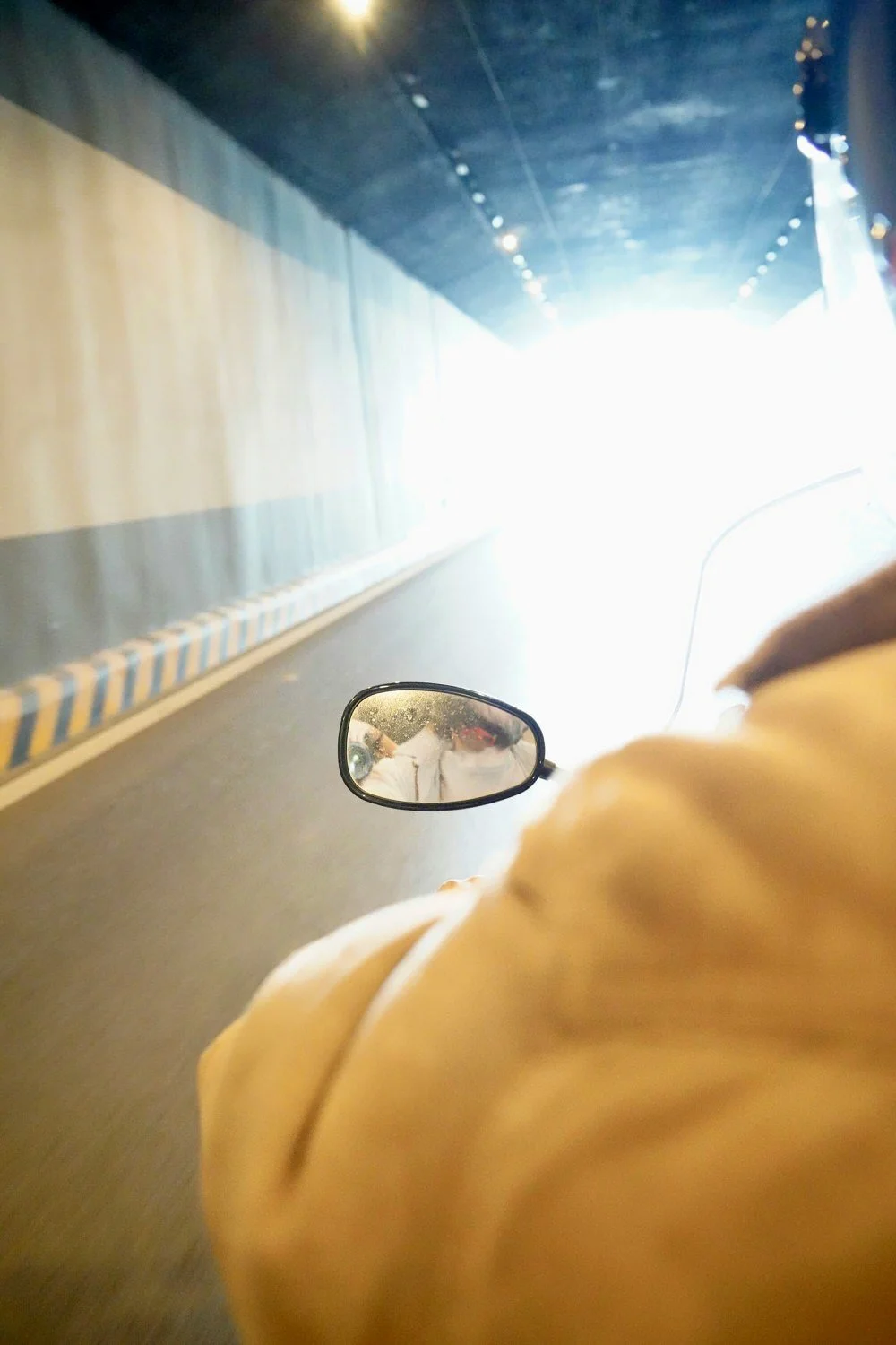 View from motorcycle rider inside a tunnel, showing the motorcycle's side mirror and rider's shoulder in a yellow jacket, with bright light at the tunnel's end.