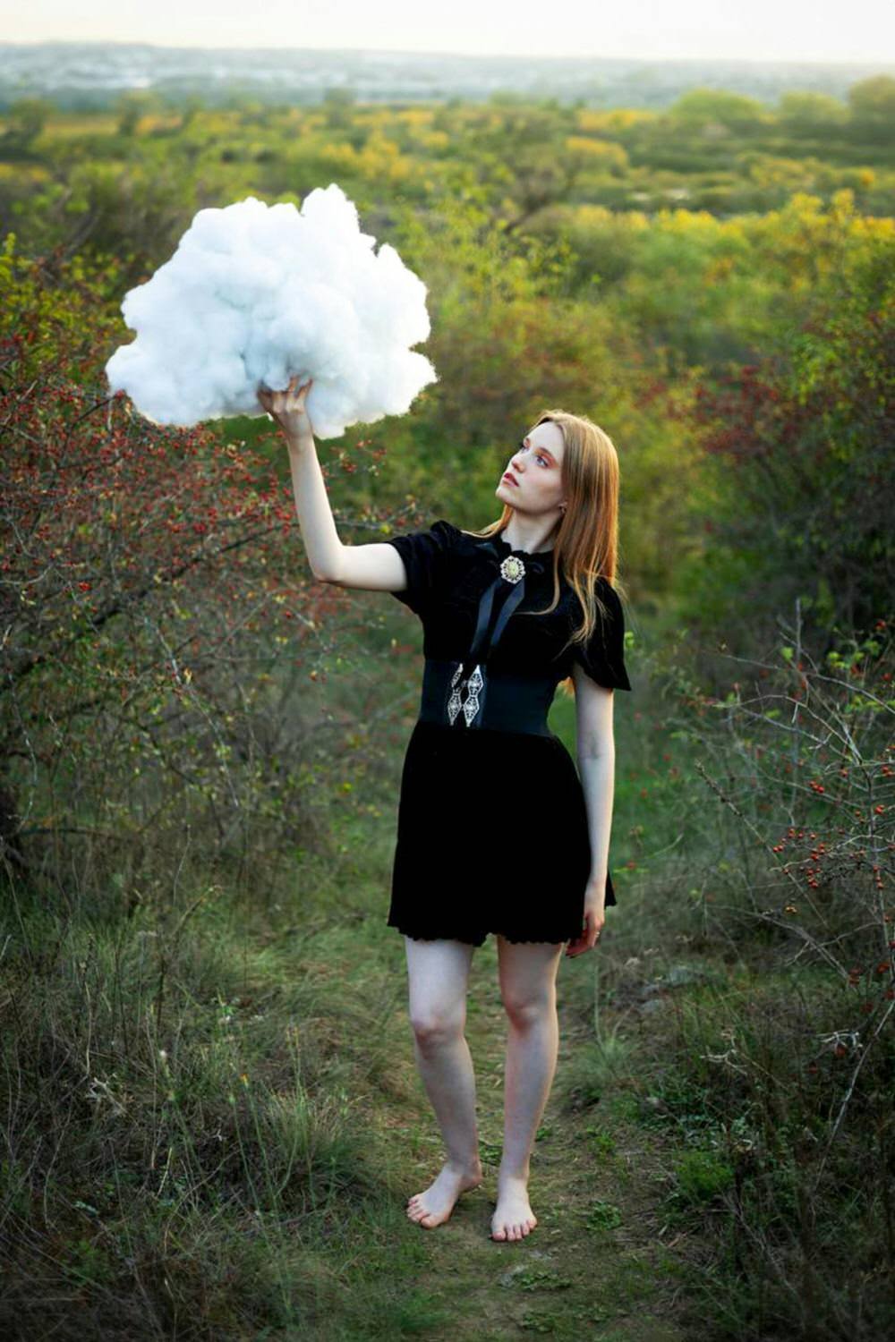 A woman with red hair standing outdoors on a dirt path, holding a large fluffy cloud-like object in her hand, surrounded by green and red foliage.
