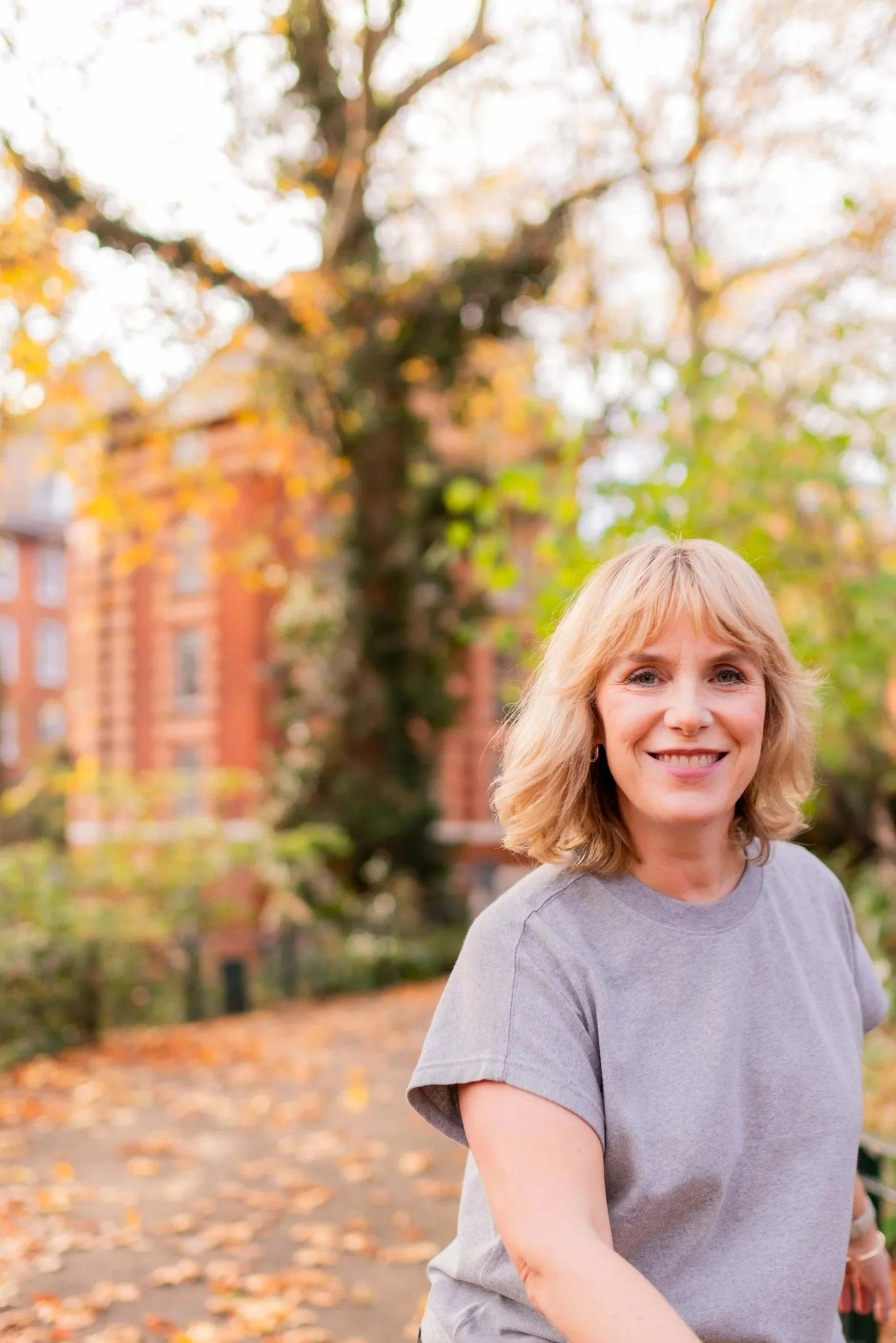 A woman with blonde hair smiling outdoors during autumn, with trees and a brick building in the background. Photo by Nicole Engelmann London Branding Photos
