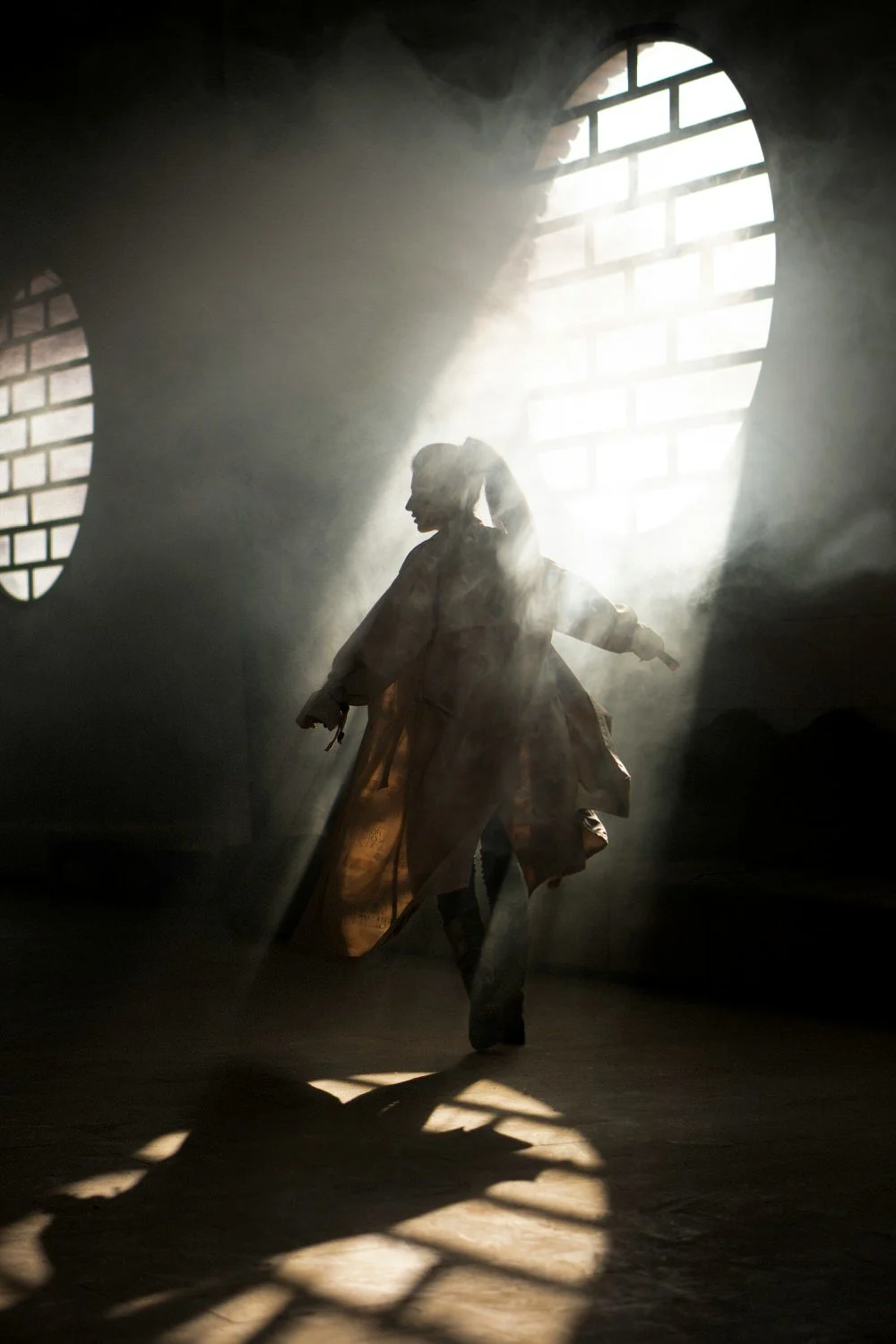 Silhouette of a person dancing in a sunlit room with large brick windows, surrounded by fog or dust.