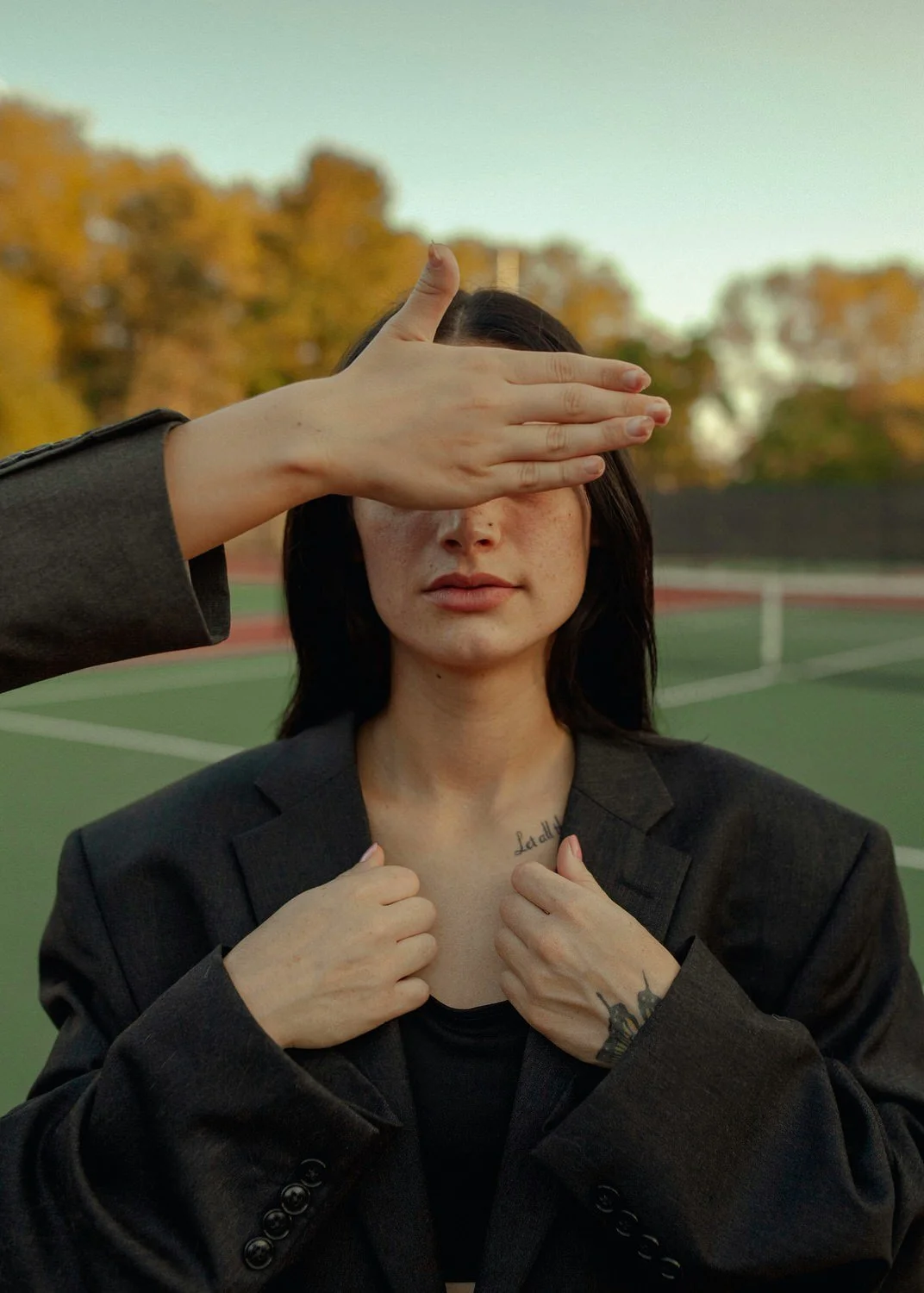 A woman with dark hair, wearing a black blazer, is being covered by a hand, which is shielding her eyes. She is outdoors on a sports field during sunset, with trees in the background.