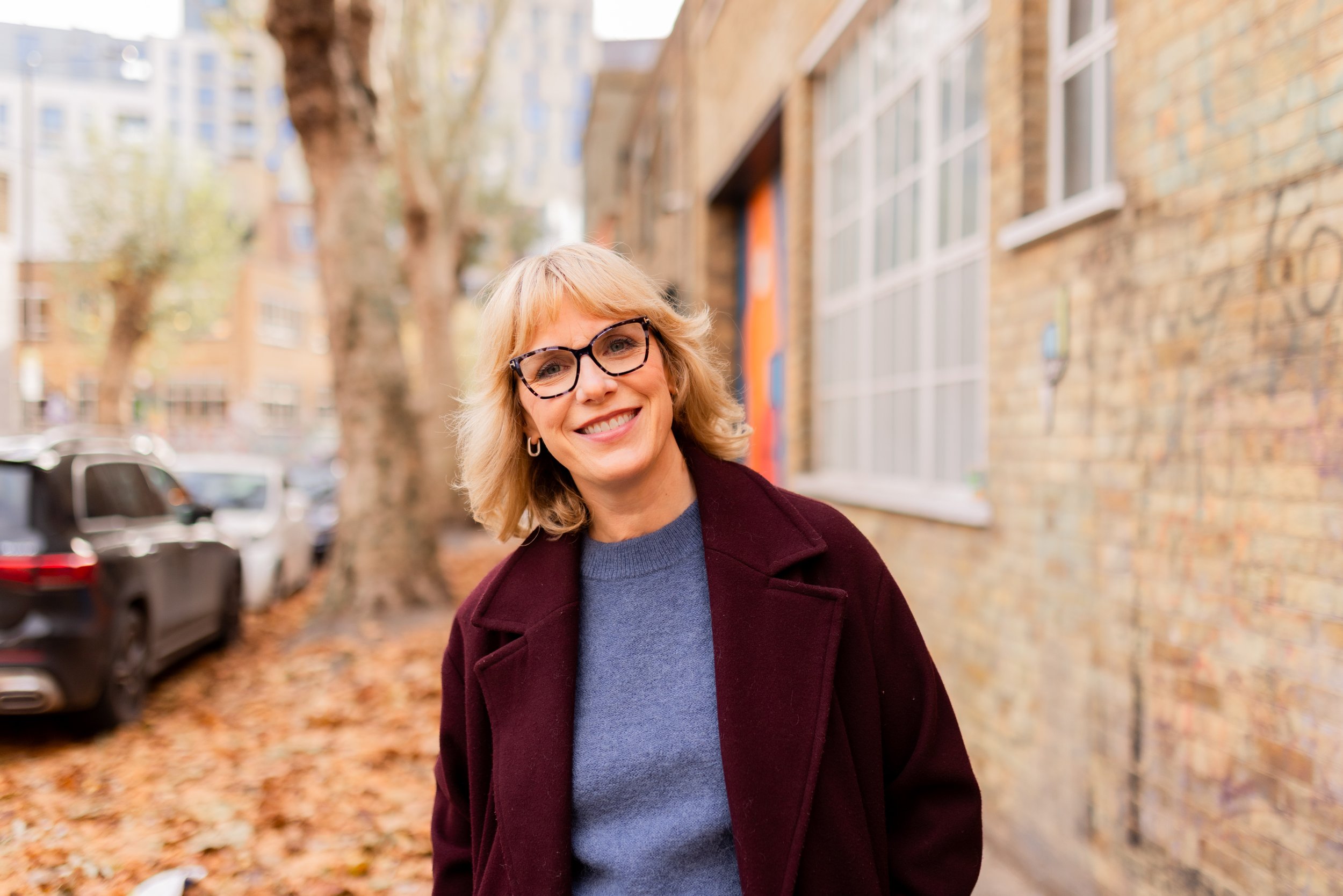 A smiling woman with blonde hair and glasses standing outdoors on a city street with fallen leaves, parked cars, trees, and brick buildings in the background.