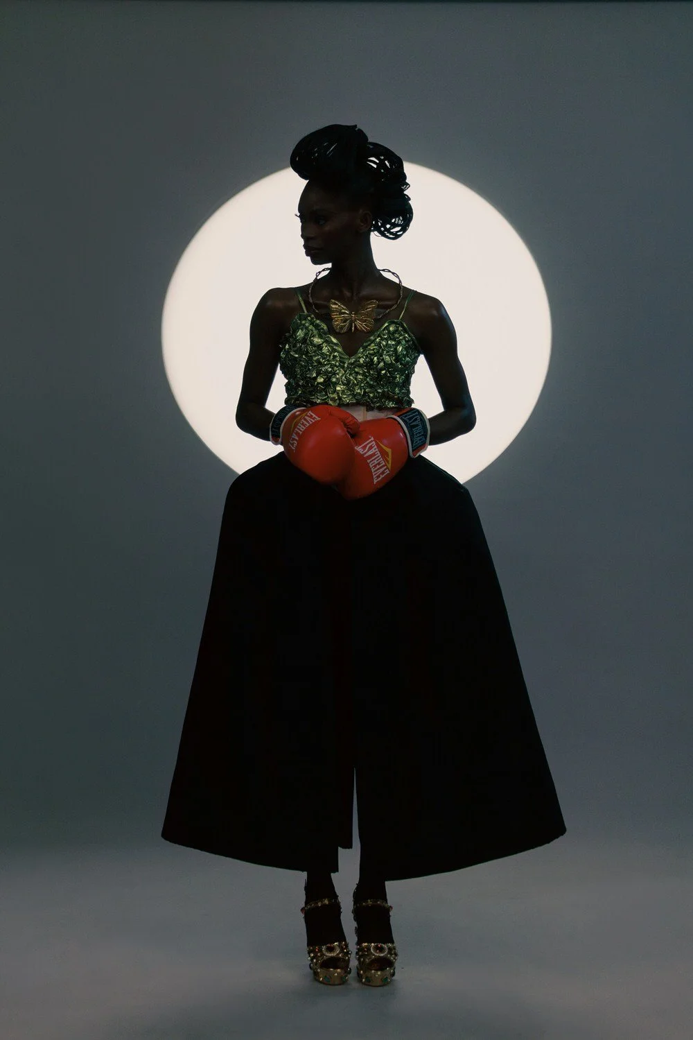 Silhouette of a woman with styled hair, wearing a shiny green top with spaghetti straps, a butterfly necklace, and flowing black wide-leg pants, holding red boxing gloves, standing in front of a circular light on a dark background.