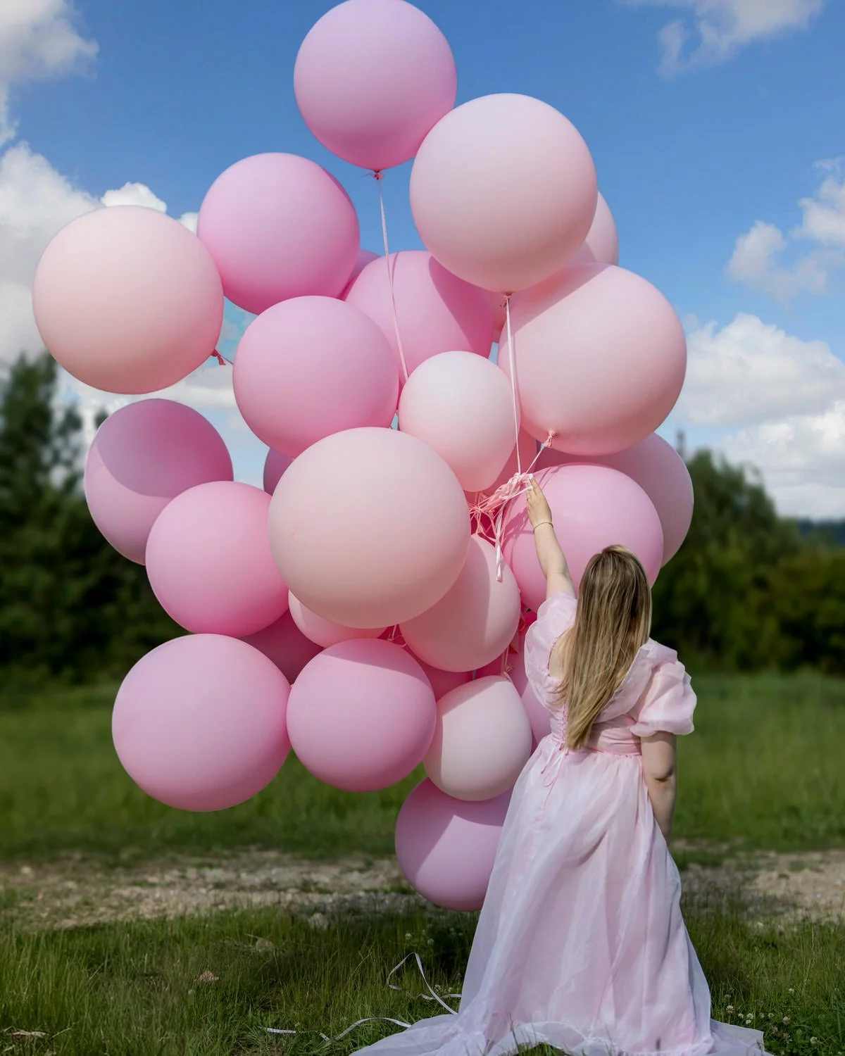 A young girl in a pink dress holding pink and white balloons outdoors on a grassy field with trees and a blue sky with clouds in the background.