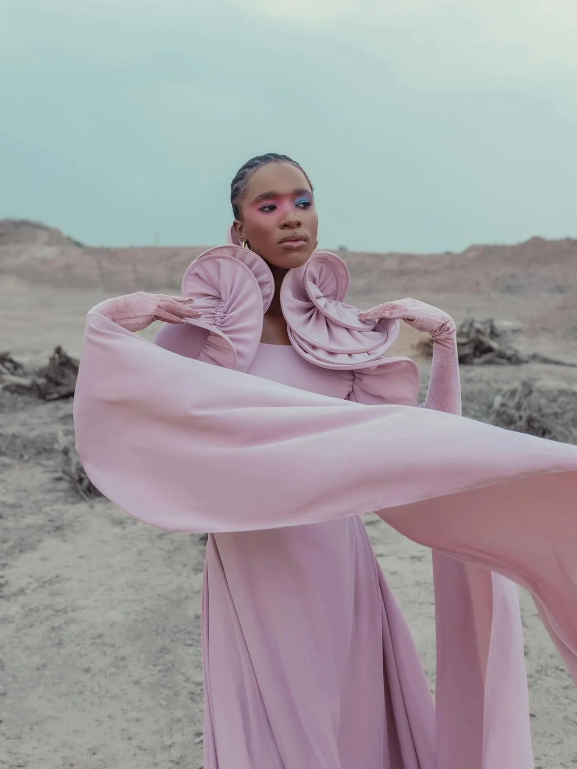 A woman in a pink, avant-garde dress with ruffled and billowing fabric, standing in a barren desert landscape with a cloudy sky.