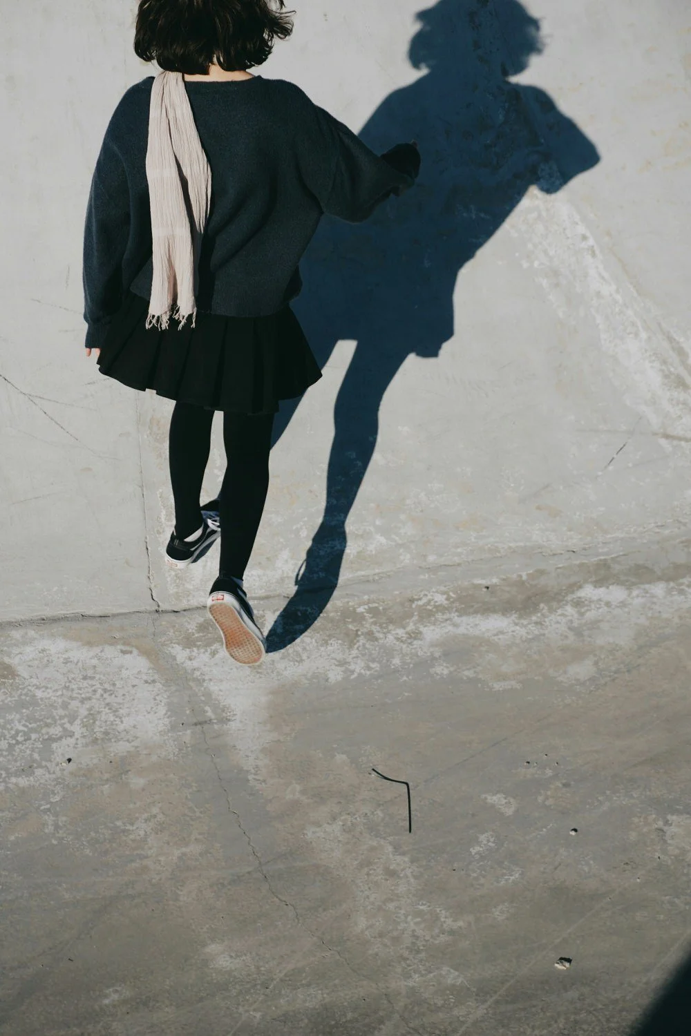 A girl with dark hair wearing a dark sweater, black pleated skirt, black tights, and sneakers casts a shadow on a concrete wall.