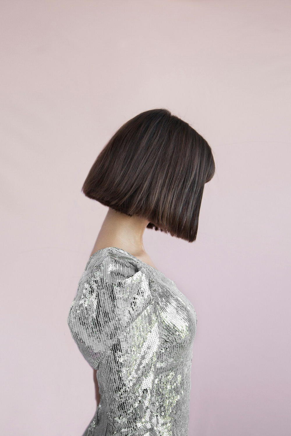 A woman with a chin-length straight bob haircut in a shiny silver sequined dress, standing sideways against a light pink background.