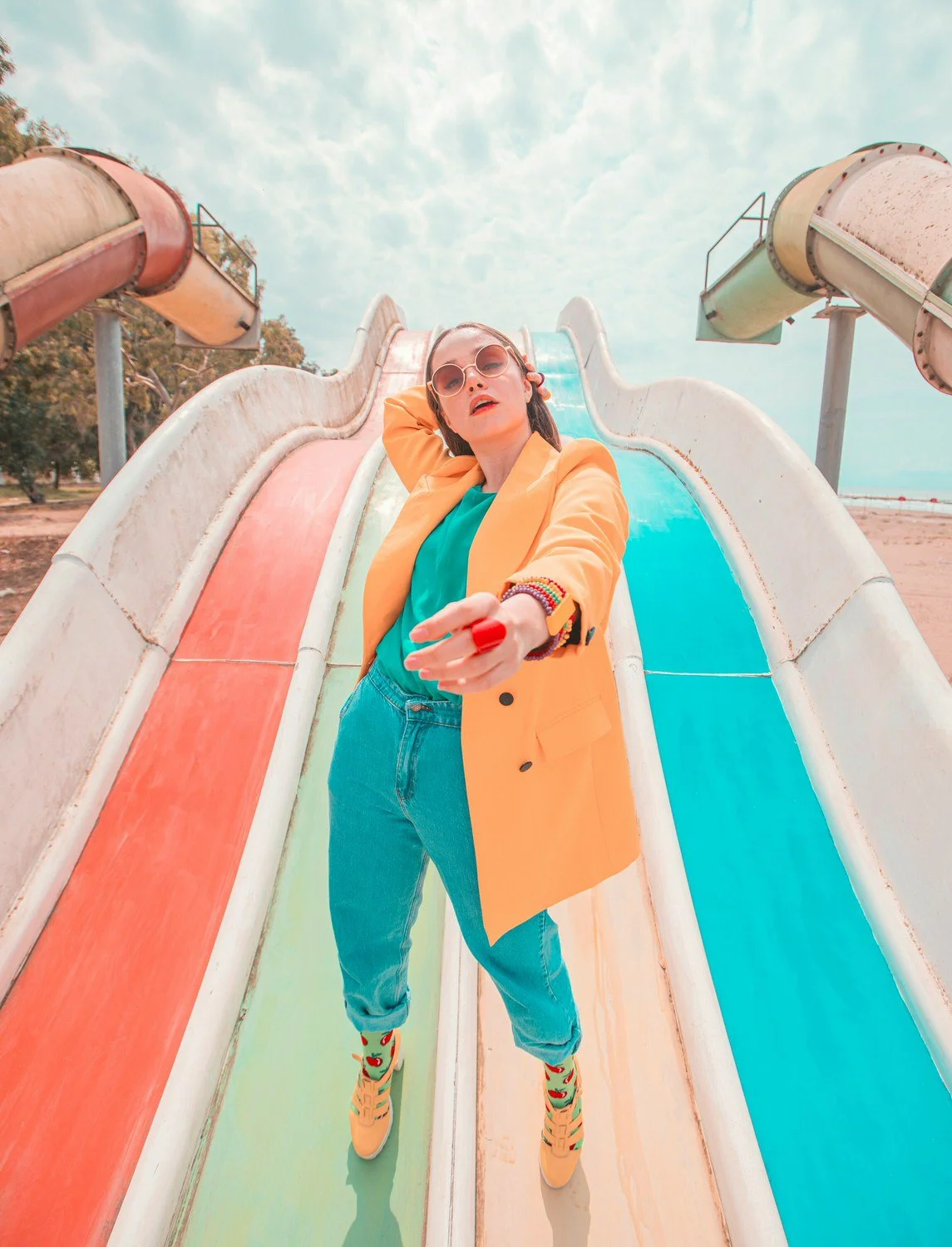 A woman wearing yellow blazer, teal shirt, jeans, and colorful socks with sneakers, posing on a rainbow-colored slide at an abandoned water park, with a partly cloudy sky in the background.
