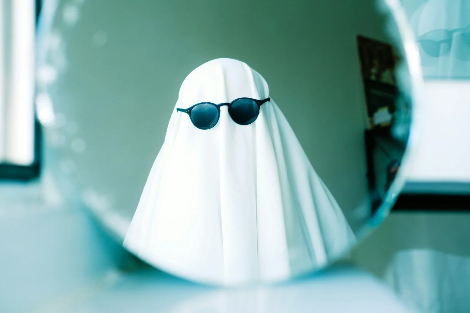 A reflection in a mirror shows a ghostly figure wearing a white sheet with sunglasses, viewed through the mirror's edge.