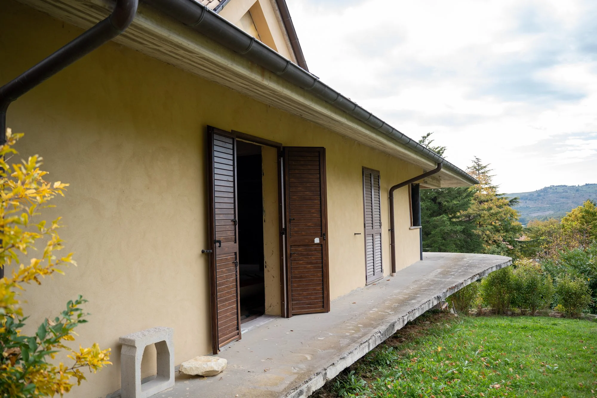 Side view of a yellow house with a balcony, brown shutters, and a view of trees and hills in the background.