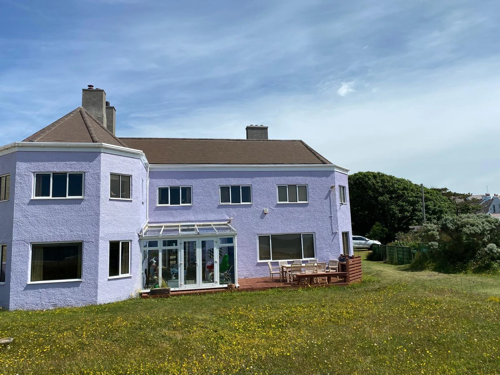 A purple three-story house with a brown roof and multiple windows, including a glass patio or sunroom, with an outdoor wooden deck and table in the backyard, and a grassy yard in front. There are trees and neighboring houses in the background under a