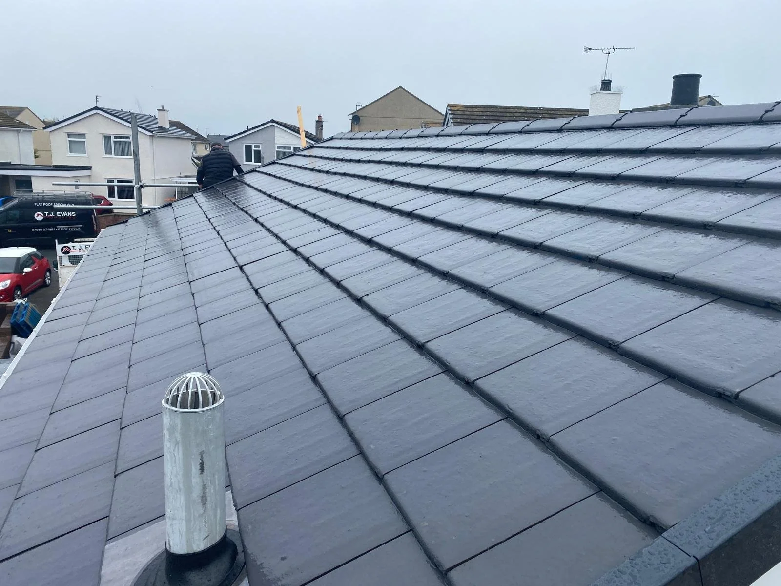 Wet tiled roof of a house with a vent pipe in the foreground and neighboring houses in the background.