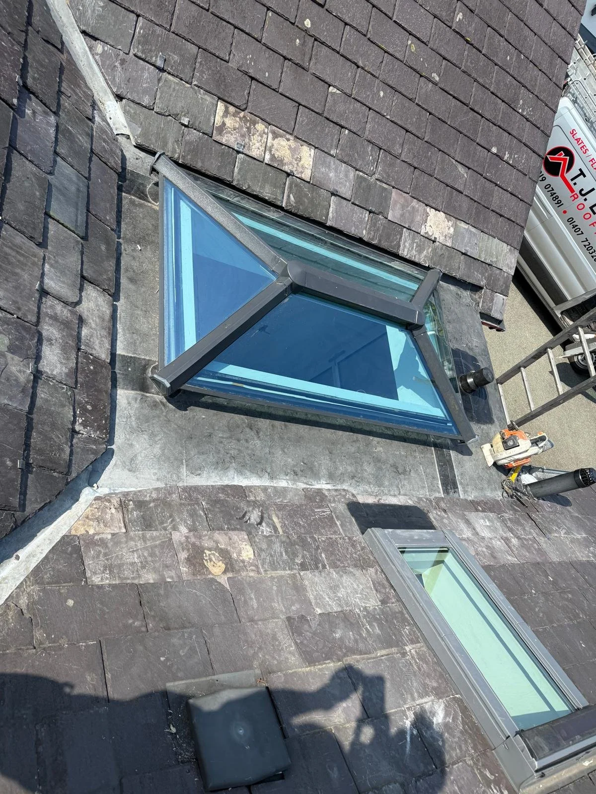 Exterior rooftop view showing a triangular skylight window on a shingled roof with some construction tools and equipment nearby.