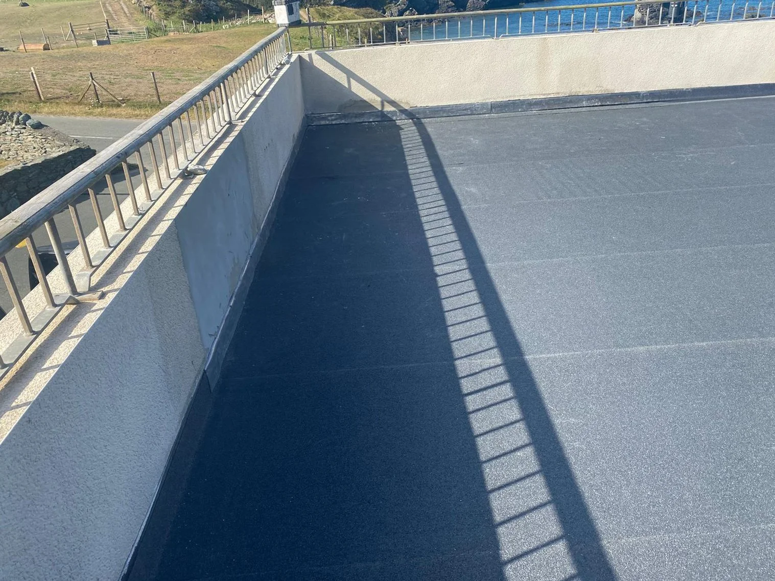 Empty rooftop with a metal railing casting a shadow on the surface, and a low wall with a patch of grassy field beyond.