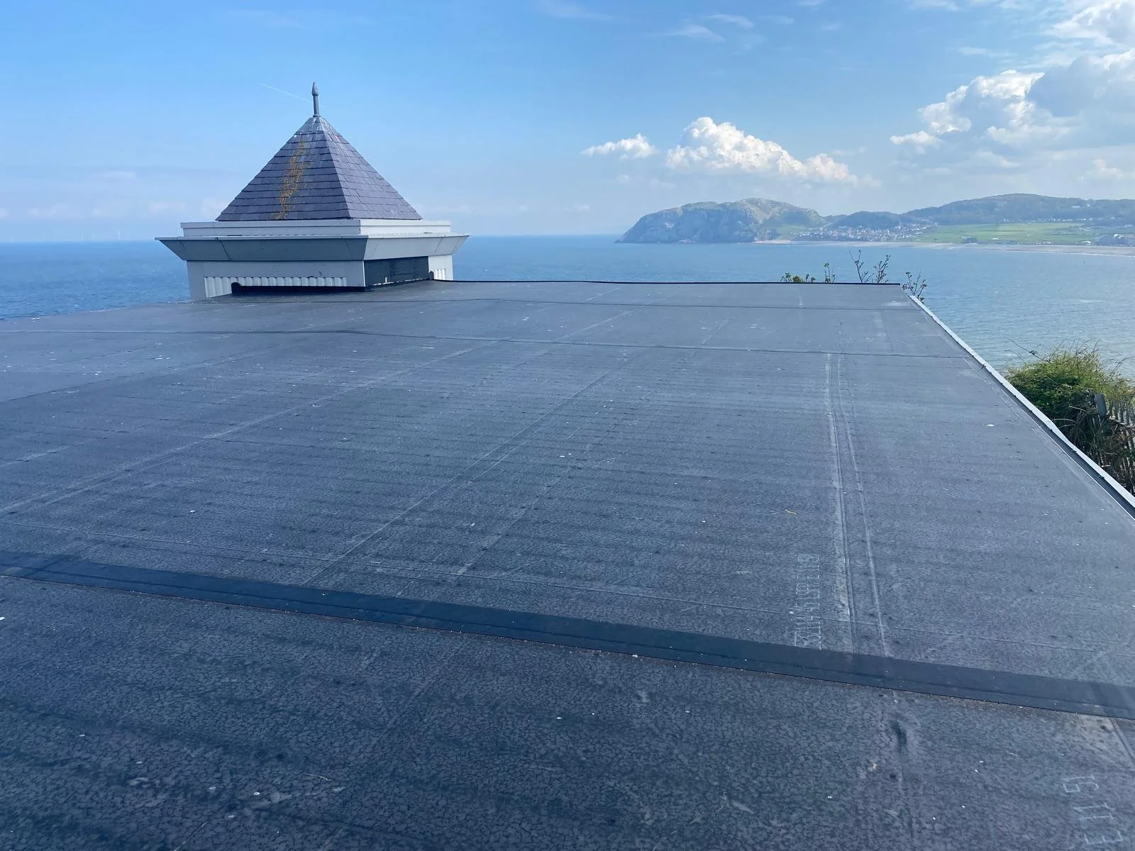 View from a rooftop with a solar panel in the foreground, a small church steeple, and the ocean with distant hills in the background.