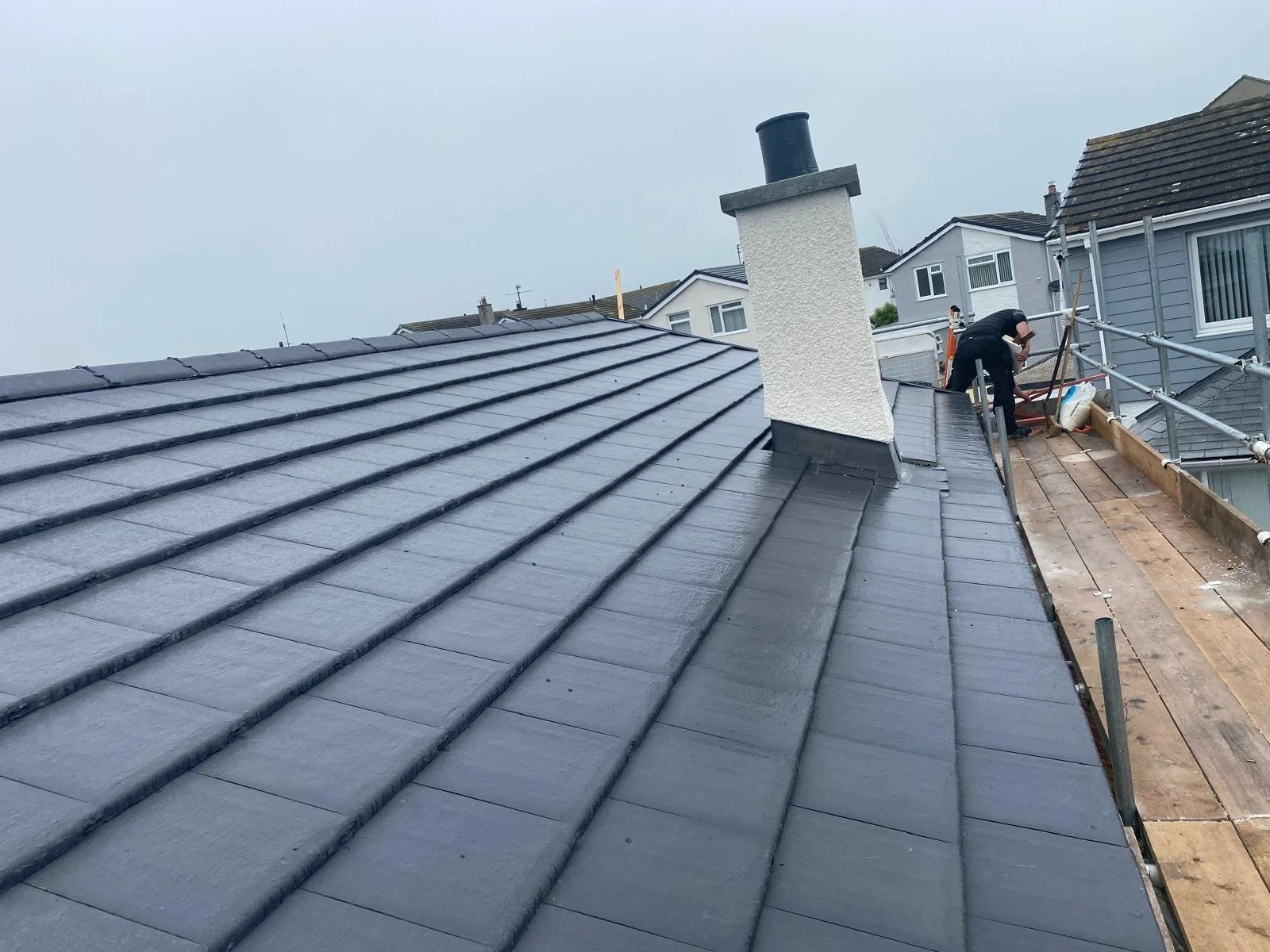 A worker installing or repairing roof shingles on a residential roof, with scaffolding and tools around.