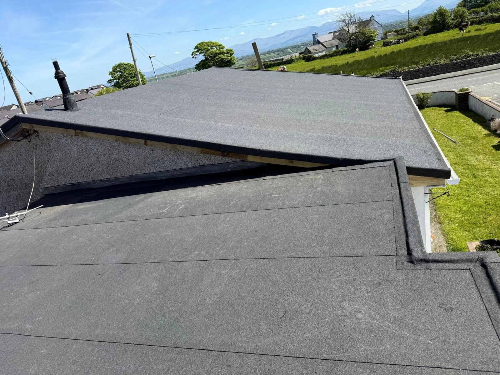 Looking down at two adjacent flat roofs covered with black gravel, with a rural landscape in the background featuring green fields, a few houses, trees, and mountains under a blue sky.