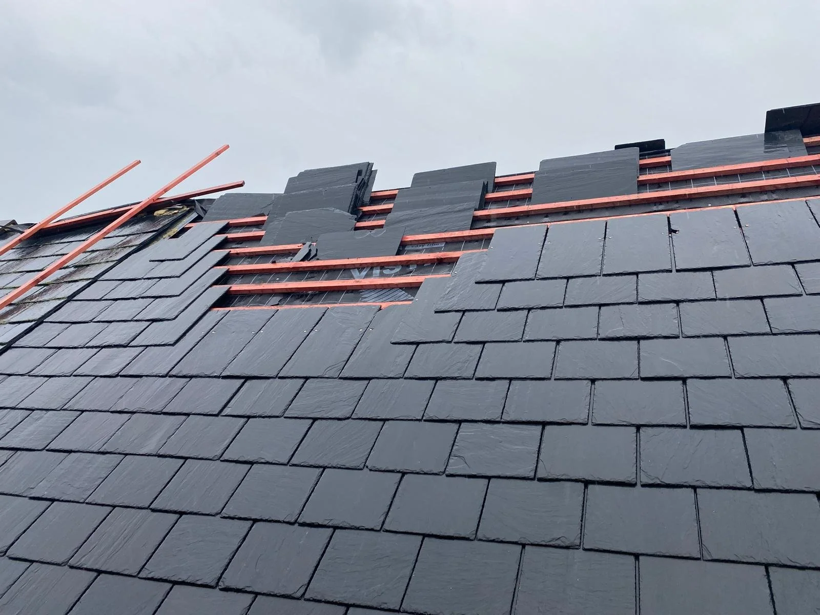 Roof under construction with black slate shingles, orange copper flashing, and exposed wooden battens on a cloudy sky background.