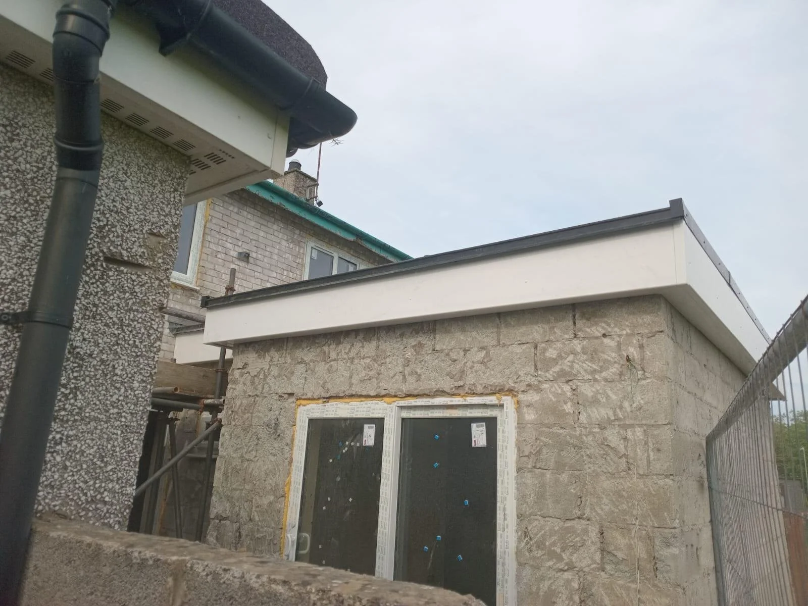Exterior view of a building under construction with stone walls, a white roof trim, a balcony, and neighboring buildings.