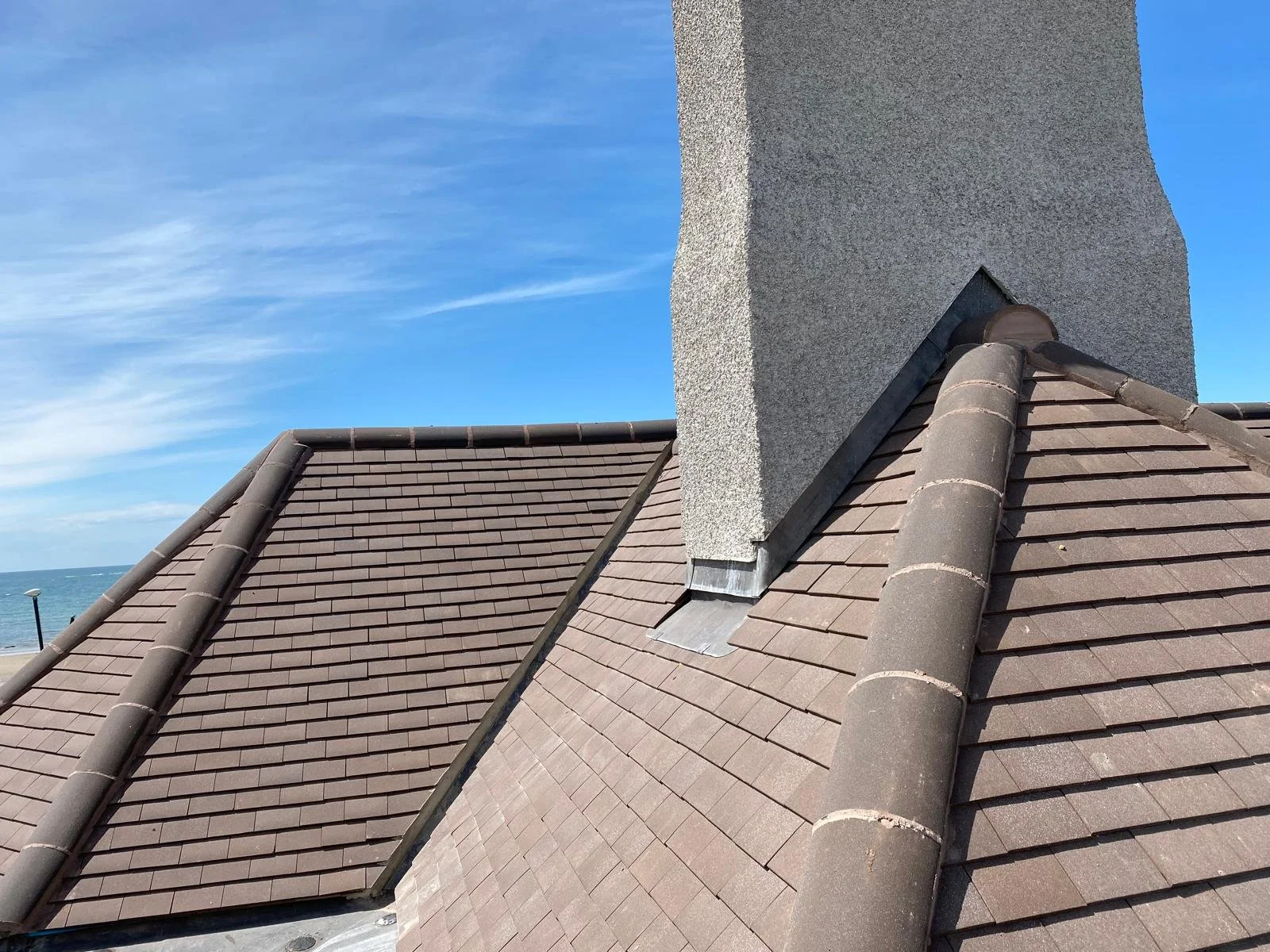 Close-up view of a roof's chimney with brown tiles and a chimney pipe, with a clear blue sky and ocean in the background.