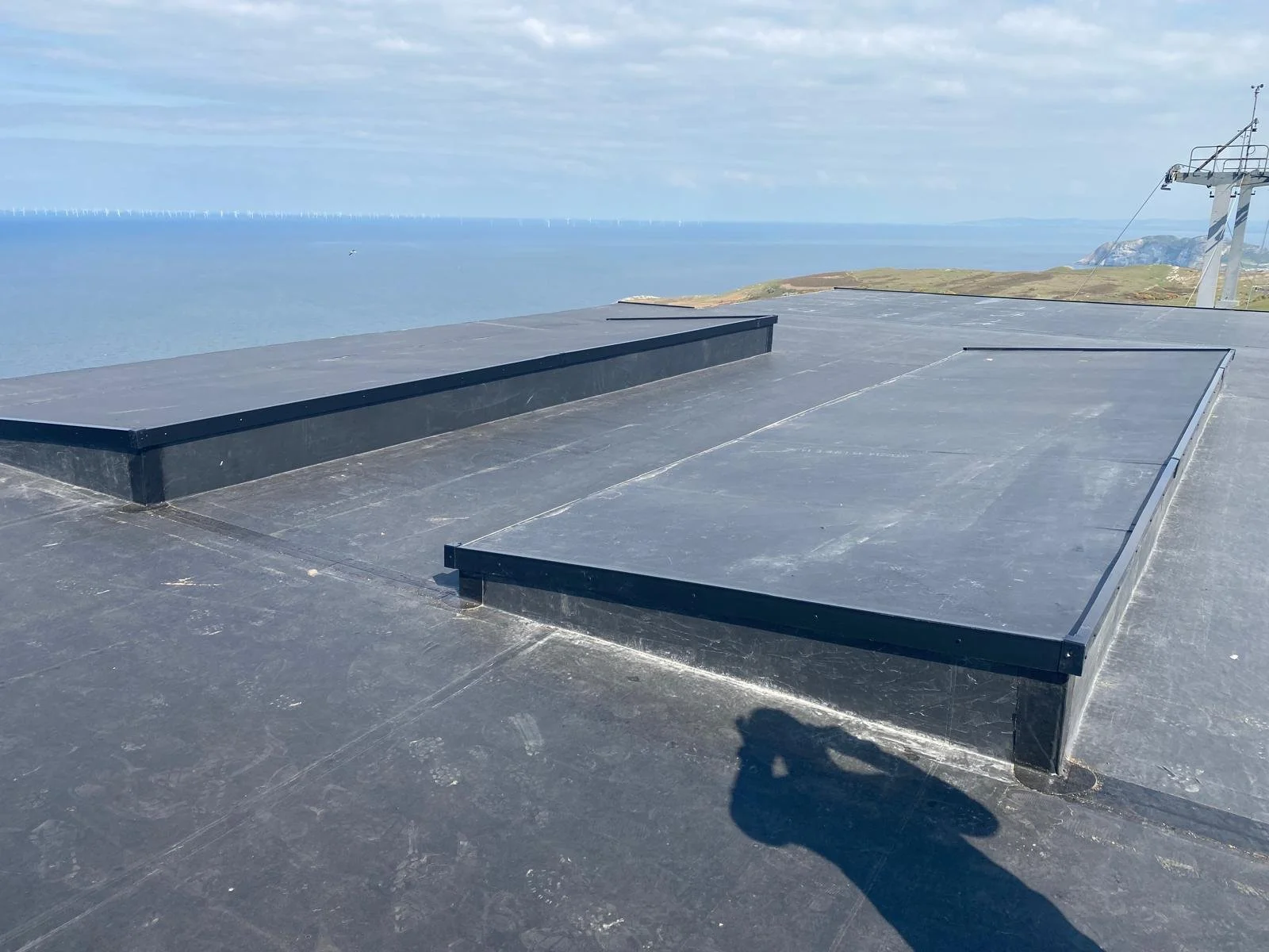 Flat, black rooftop with skylights, overlooking the ocean and cloudy sky, in a coastal area.