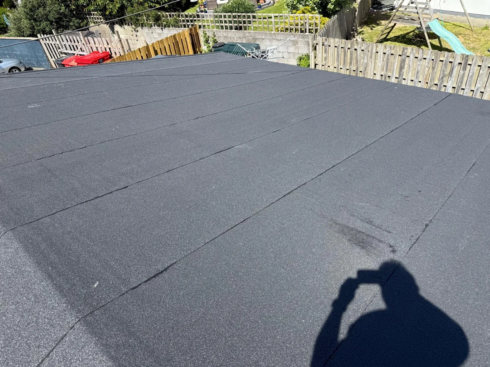 Black asphalt roof with visible seams and a person's shadow holding a camera, in a backyard setting with fences, trees, and a slide in the background.