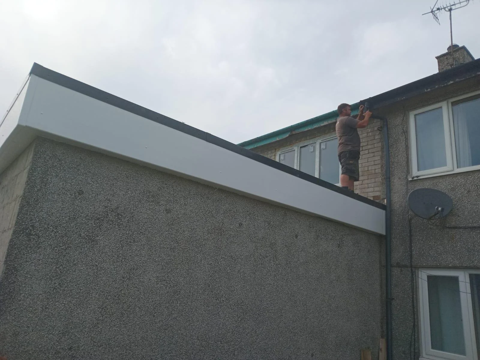 A man standing on the roof of a building, working on the gutter near the chimney, with a satellite dish attached to the exterior wall.