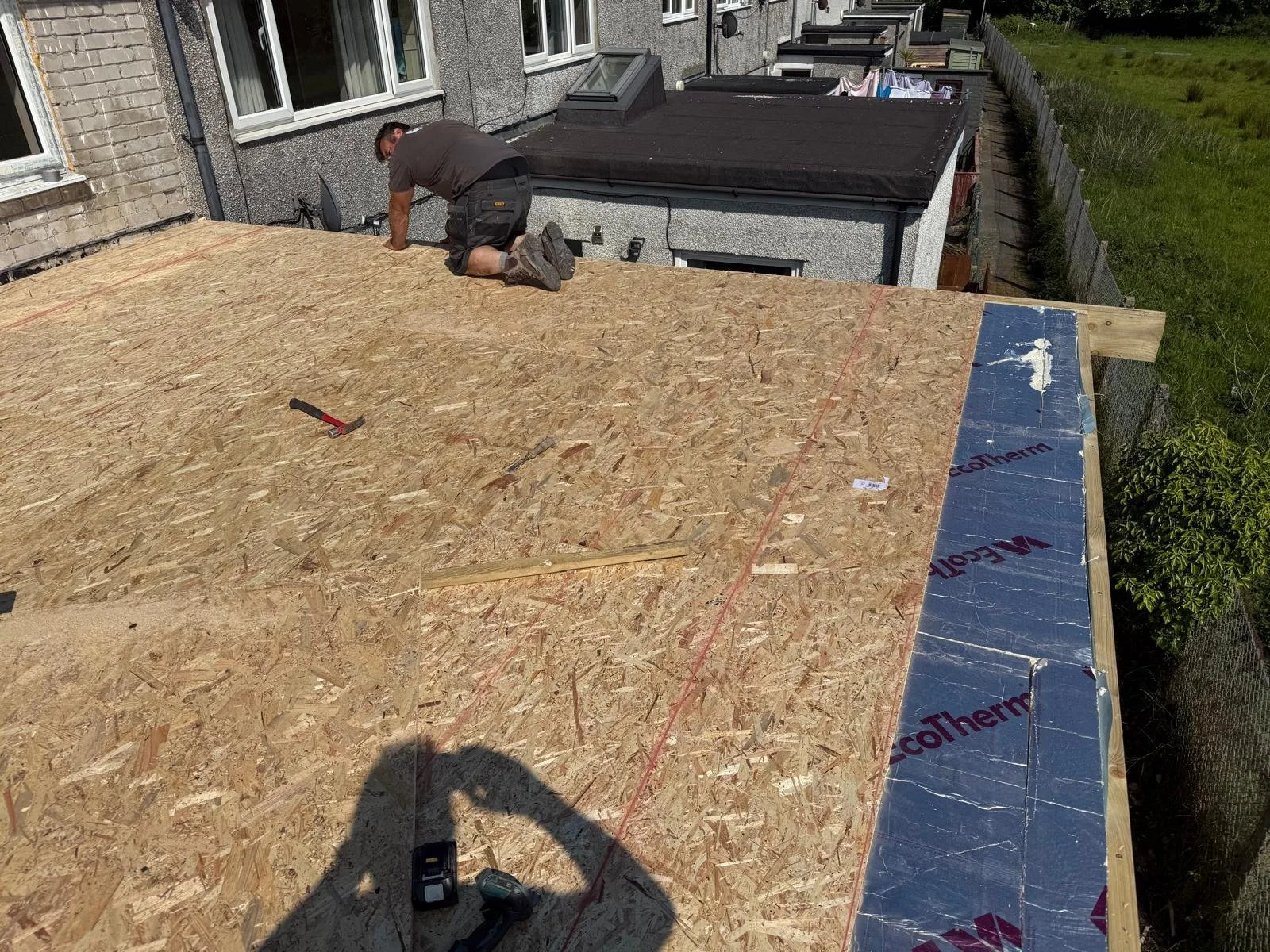 A man is working on a flat rooftop with plywood sheathing, surrounded by construction tools, with neighboring houses and a grassy area in the background.