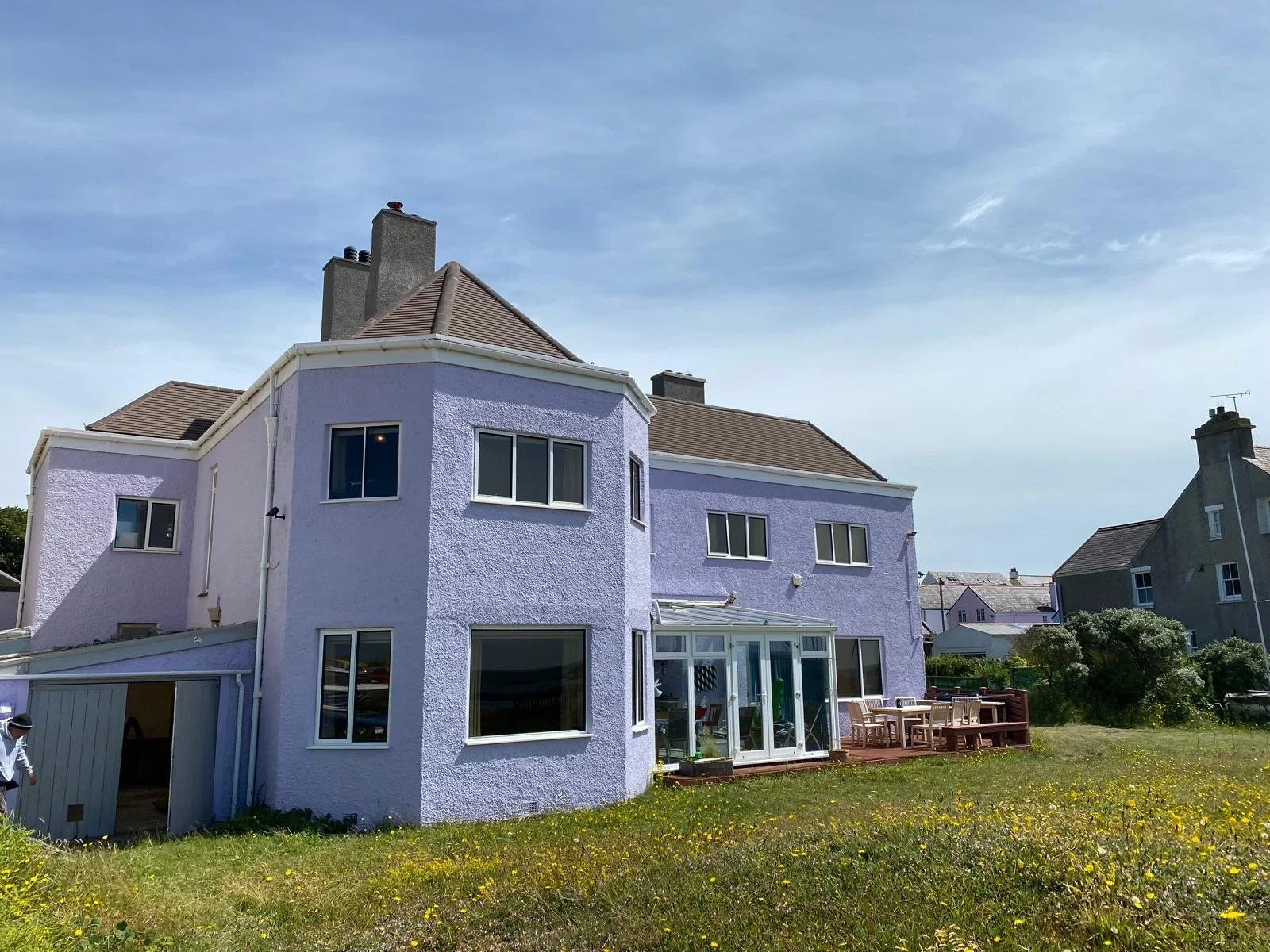 A purple multi-story house with large windows, a glass-enclosed porch, and a wooden deck in a grassy yard with yellow flowers, under a blue sky with some clouds.