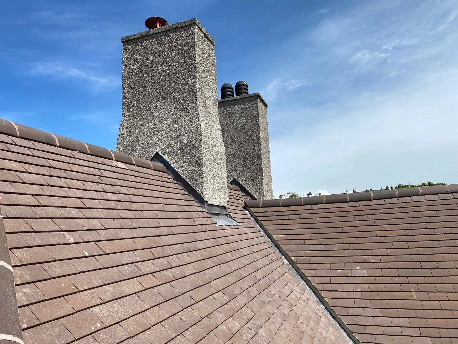 View of a rooftop with brown tiles and two large chimneys against a blue sky with some clouds.