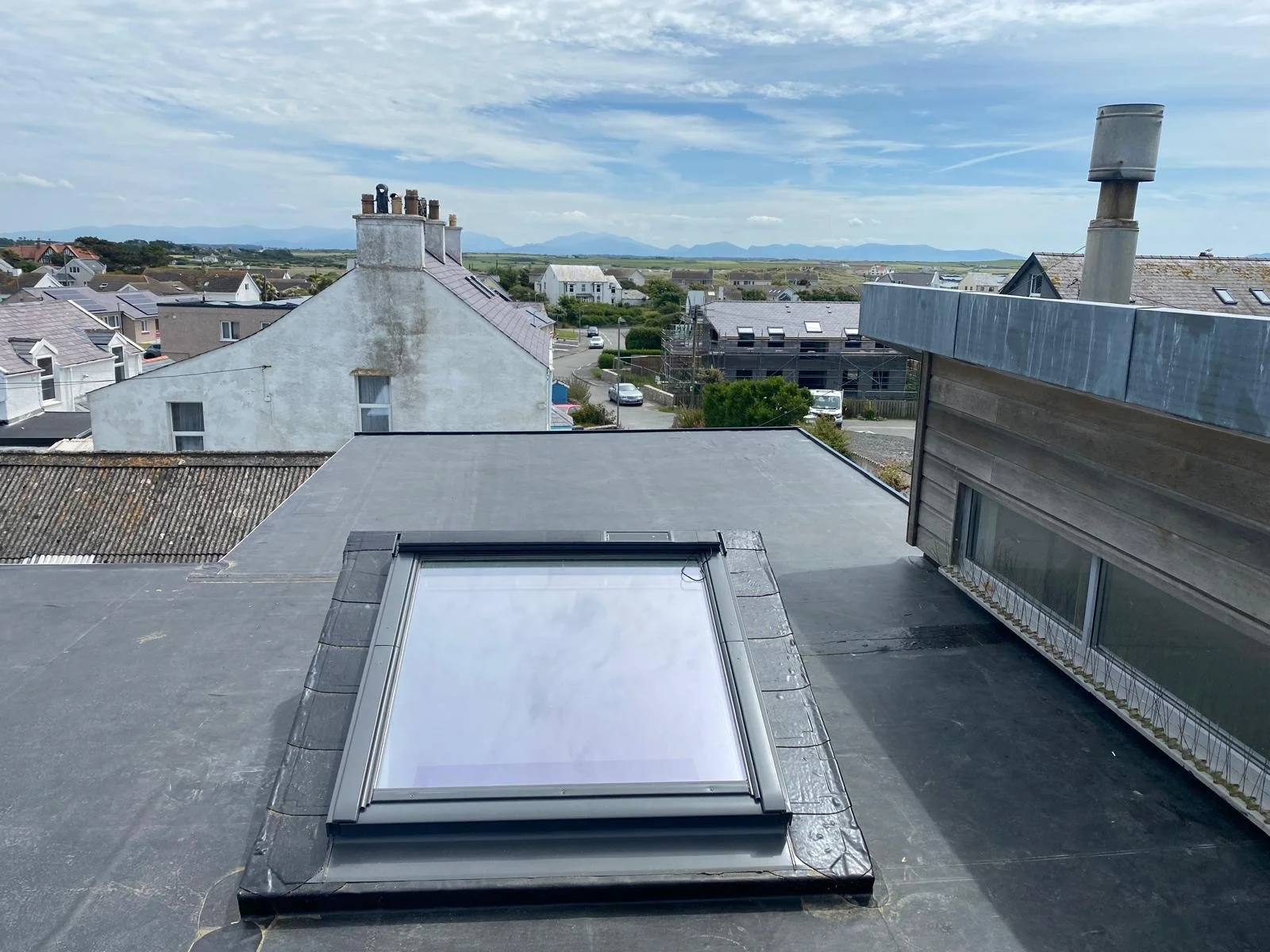 View from a rooftop showing a skylight, neighboring rooftops, and a distant landscape with mountains under a partly cloudy sky.