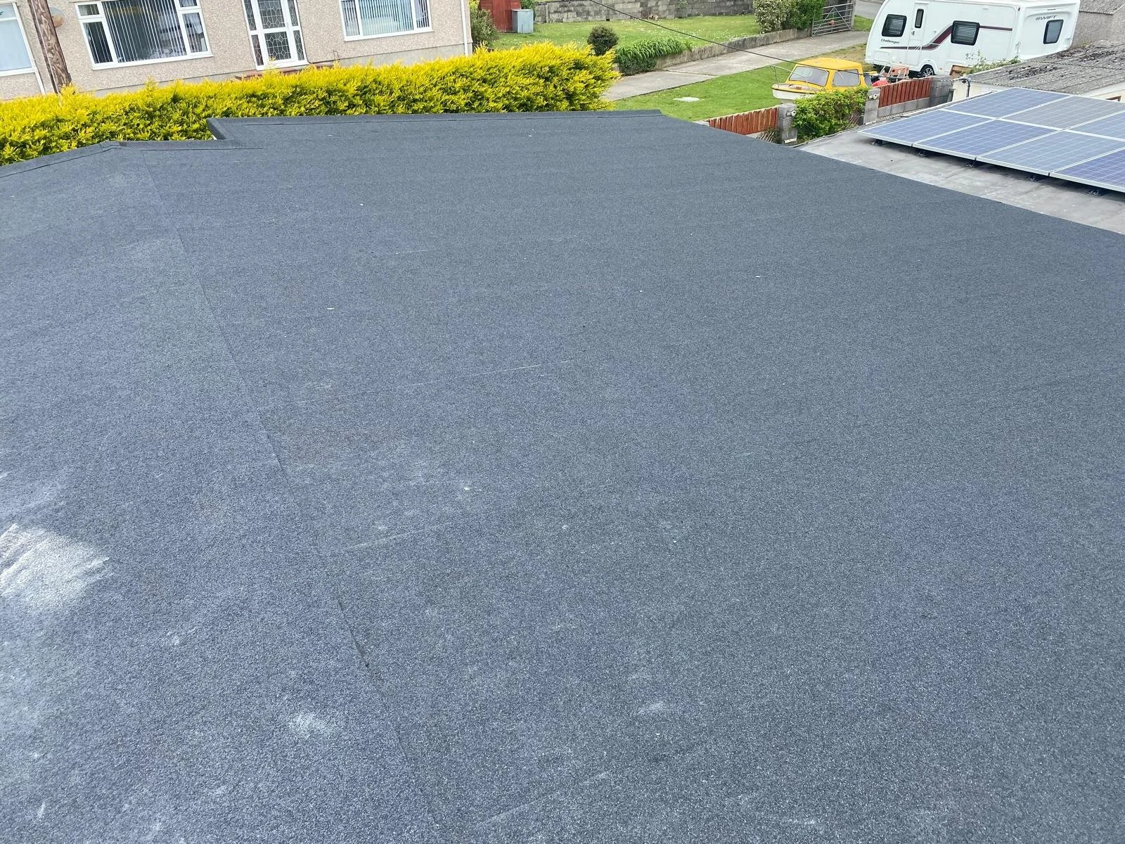 Flat roof with black asphalt shingles, with neighboring houses, trees, and solar panels in the background.