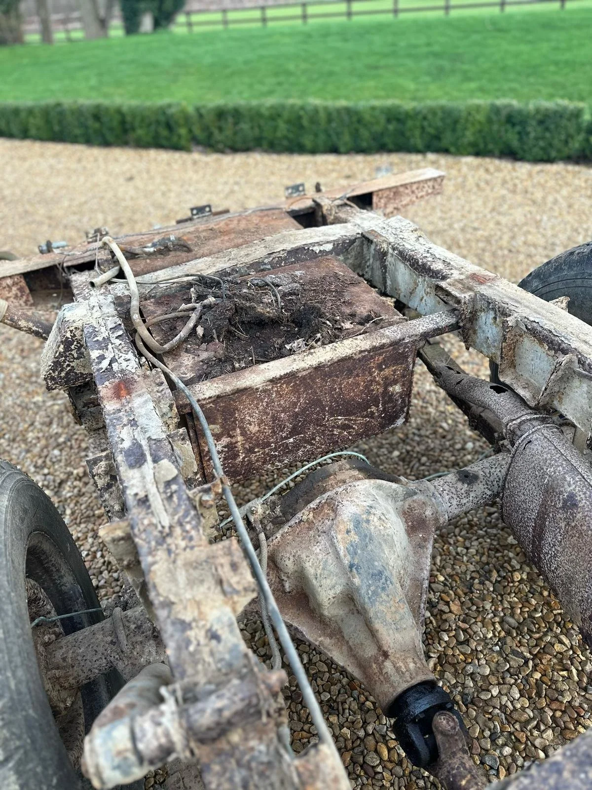 Rusty old vehicle chassis with exposed wheels and differential, parked on gravel with green lawn and hedges in the background.
