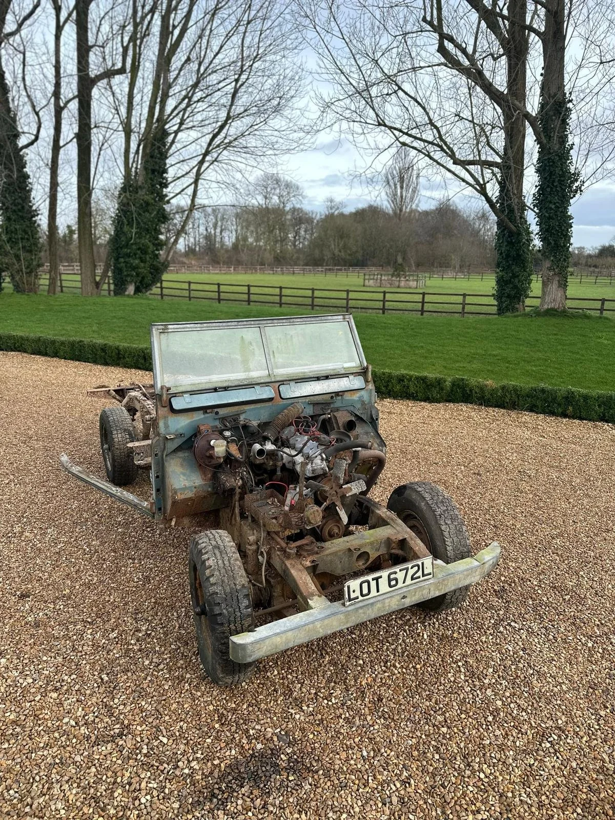 An old, rusty, disassembled car with no doors or engine parts in a rural outdoor setting on gravel, with a background of green grass, trees, and wooden fences under an overcast sky.