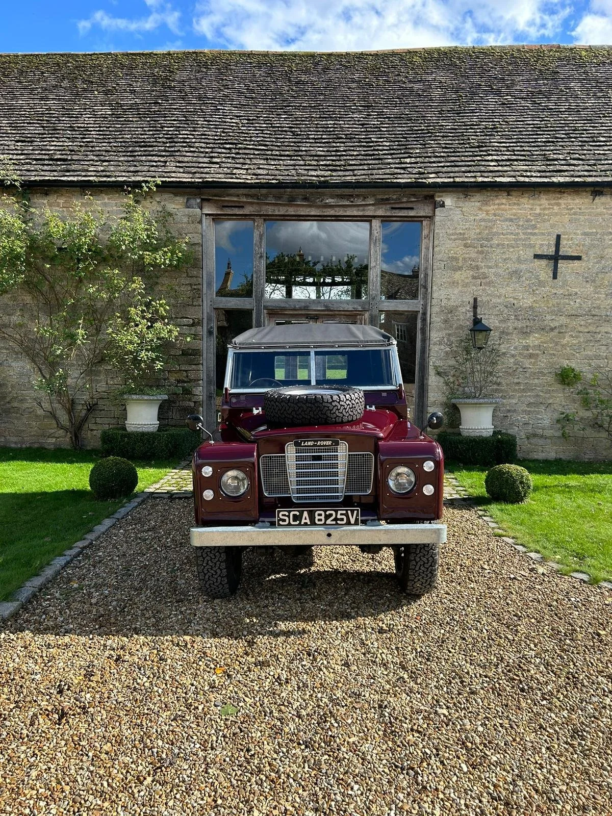 A vintage maroon Land Rover parked on a gravel driveway in front of an old stone barn with large windows, flanked by two large white planters and small topiary shrubs, under a blue sky with clouds.