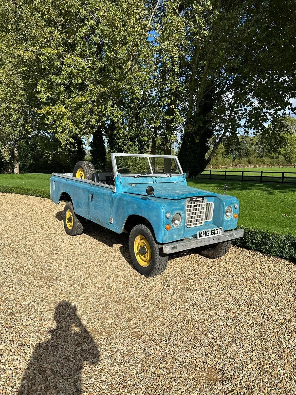 A vintage blue Land Rover with yellow wheels parked on a gravel driveway in front of green grass and trees.