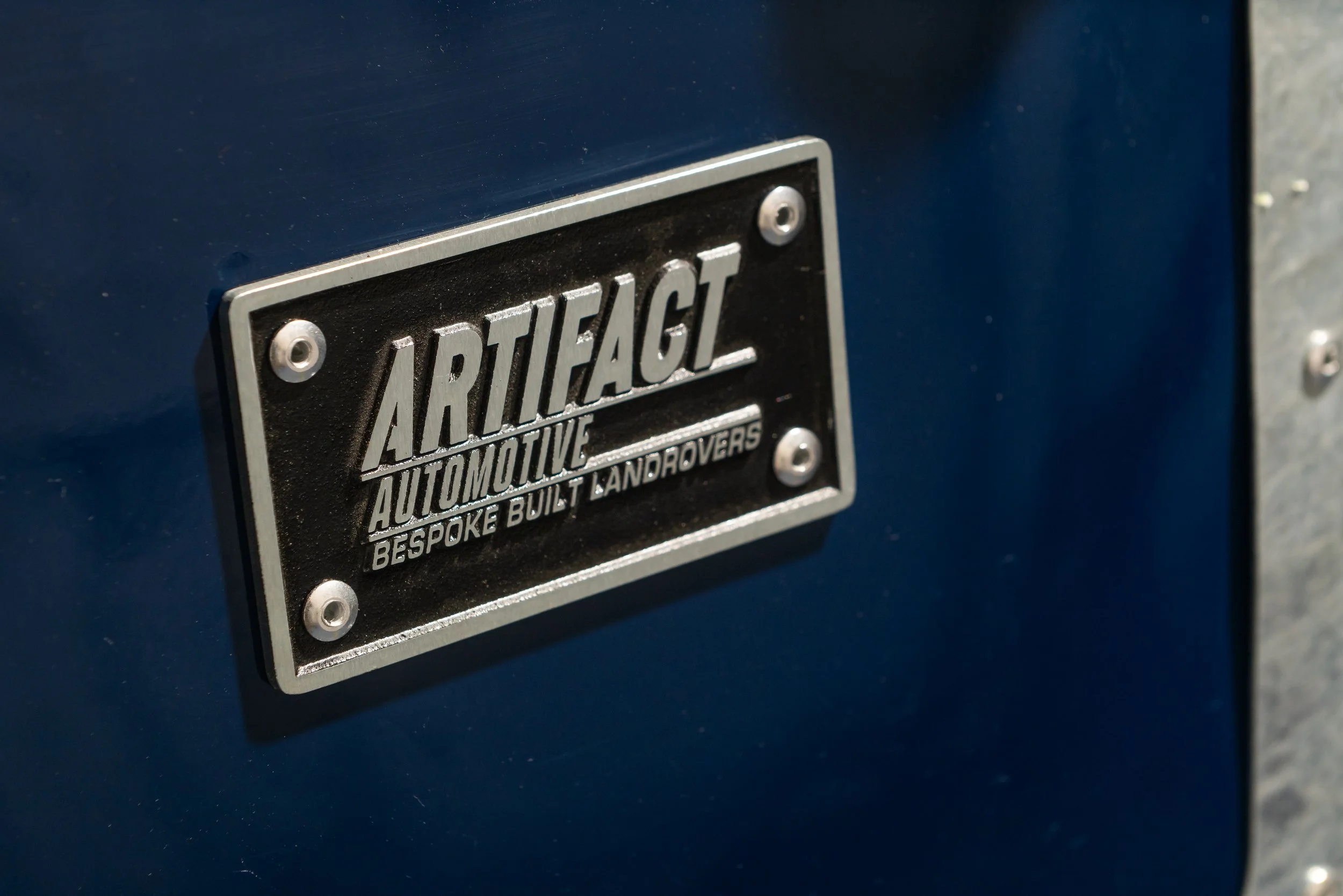 Close-up of a black and silver plate on a blue vehicle that reads 'Artifact Automotive, Bespoke Built Landrovers'.