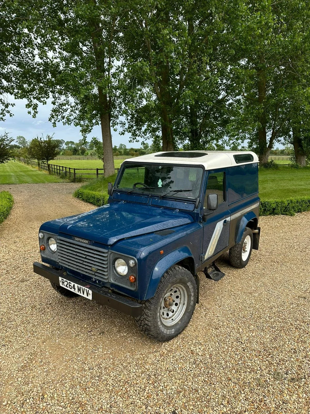A blue vintage Land Rover Defender parked on a gravel driveway in a rural setting with green trees and a field in the background.