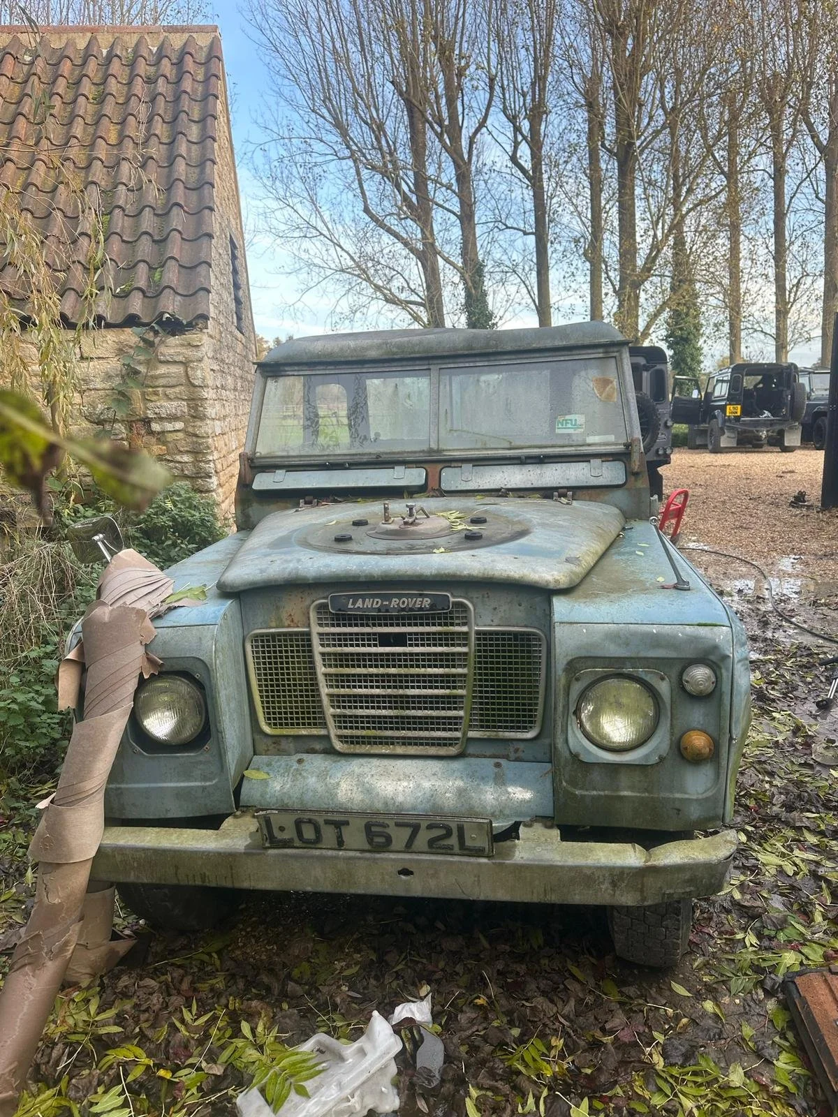 Old, rusty Land Rover parked outdoors surrounded by fallen leaves, with a brick building and leafless trees in the background.