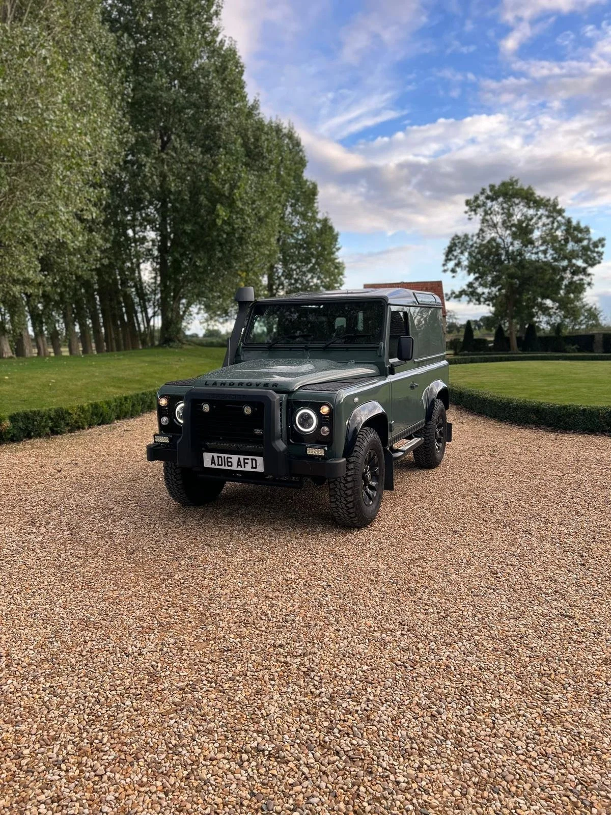 A black Land Rover Defender parked on a gravel driveway, with trees and a grassy area in the background under a partly cloudy sky.