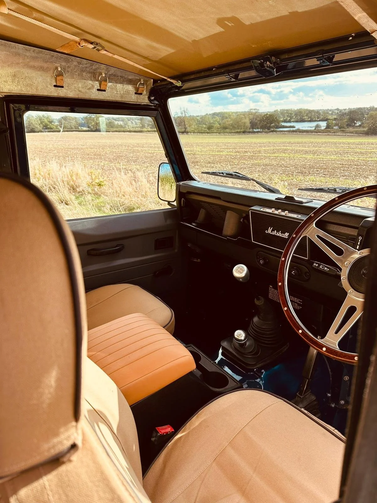 Interior of a vintage vehicle showing a beige and orange seat, a wooden steering wheel, and a dashboard with a Marshall speaker, with a view of a rural landscape through the windshield.